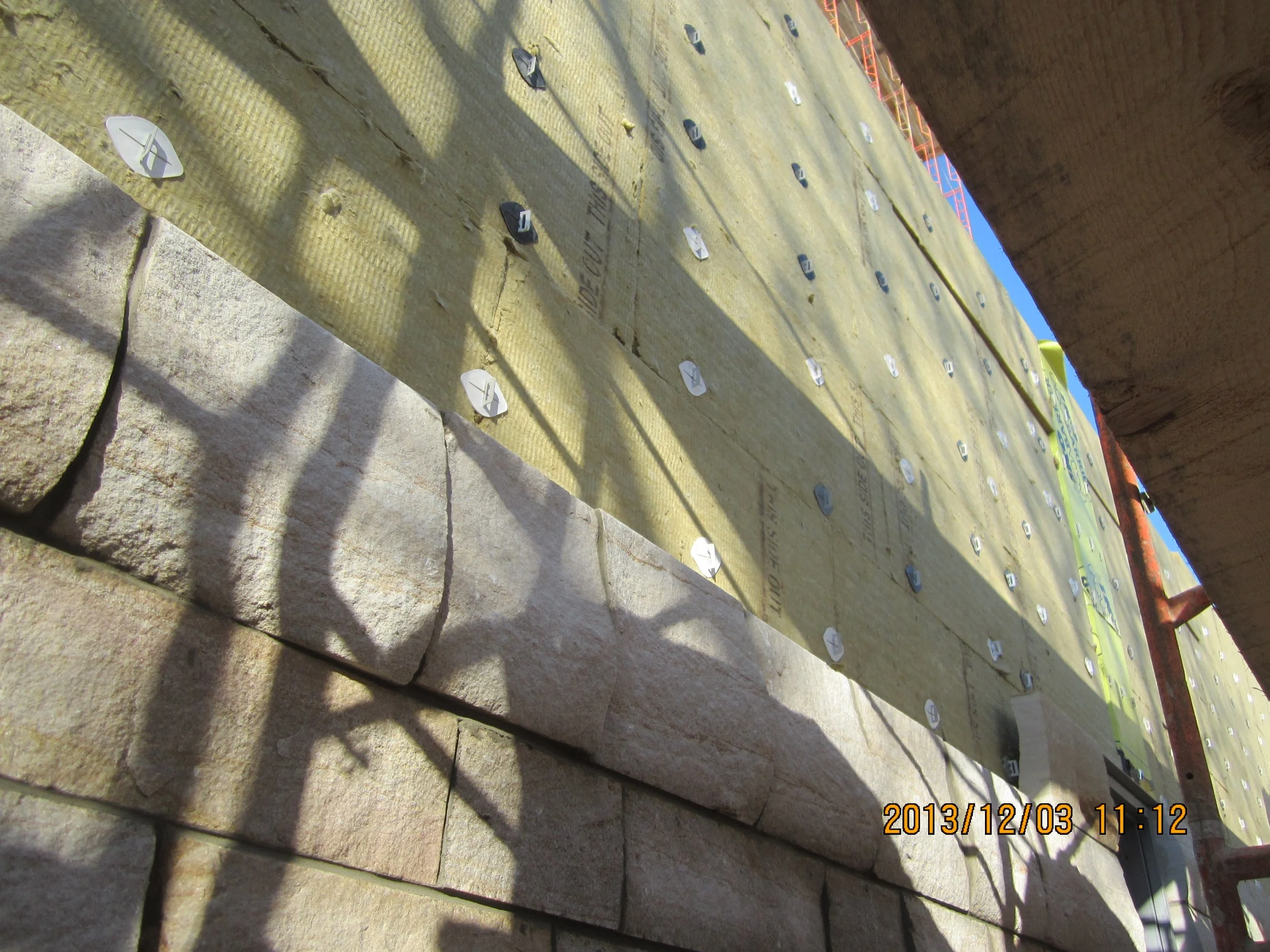 Close-up of a construction site showing a brick wall partially built, with scaffolding and insulation material on the upper wall with metal fasteners. Shadows cast on the brick wall and insulation material with a timestamp on the lower corner reading