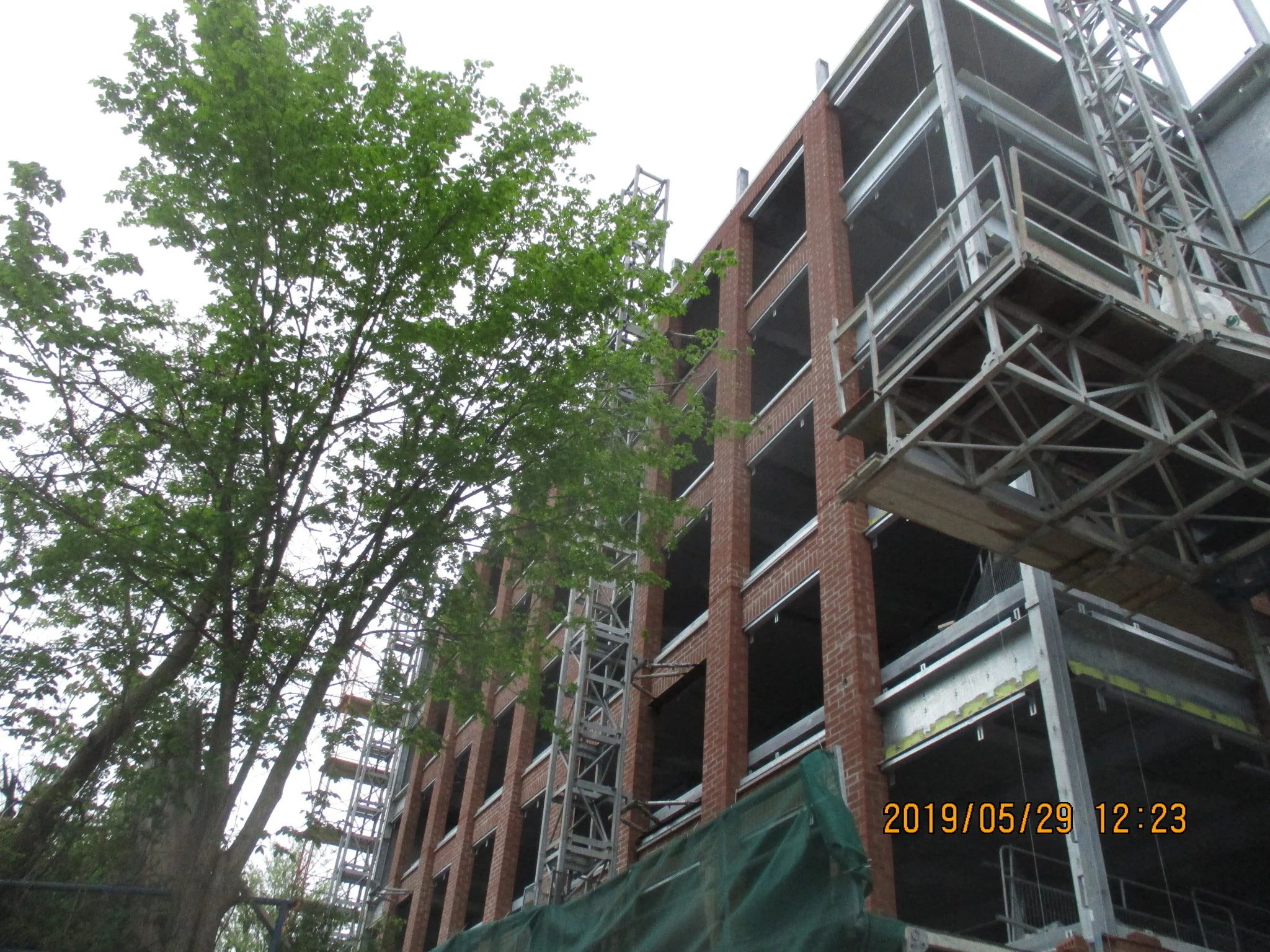 Construction site of a multi-story building with brick walls and steel framework, with a tree partially blocking the view, and a timestamp indicating the photo was taken on May 29, 2019, at 12:23 PM.