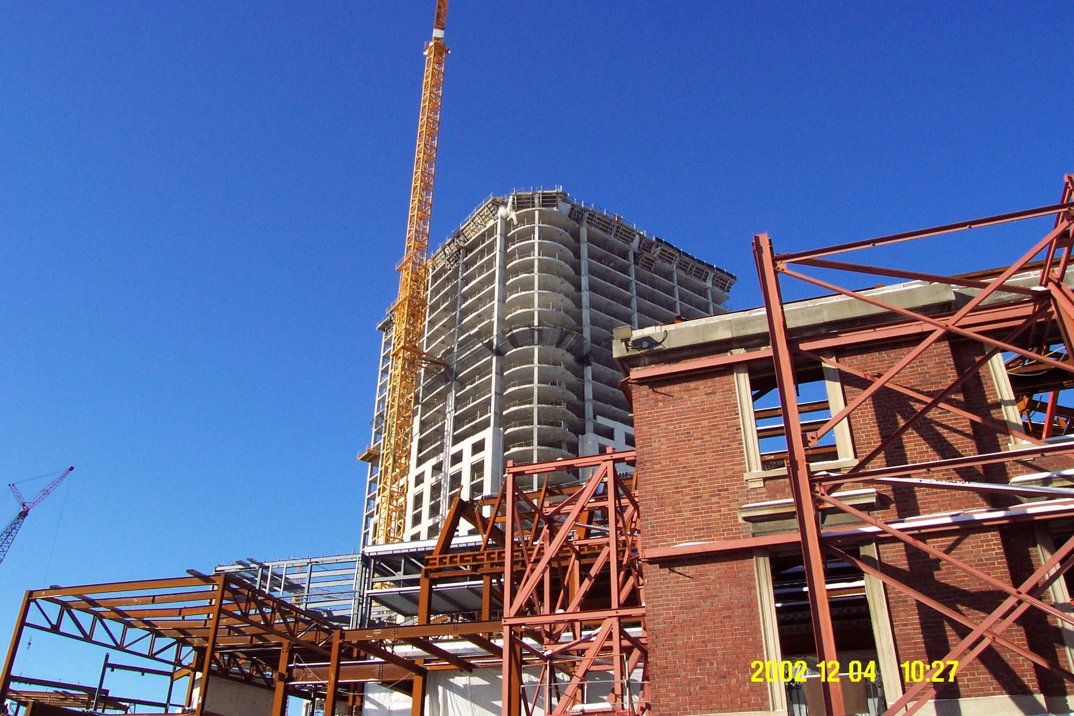 A high-rise building under construction with a yellow tower crane beside it, and a brick building in the foreground against a clear blue sky. The photo is timestamped December 4, 2002, at 10:27.