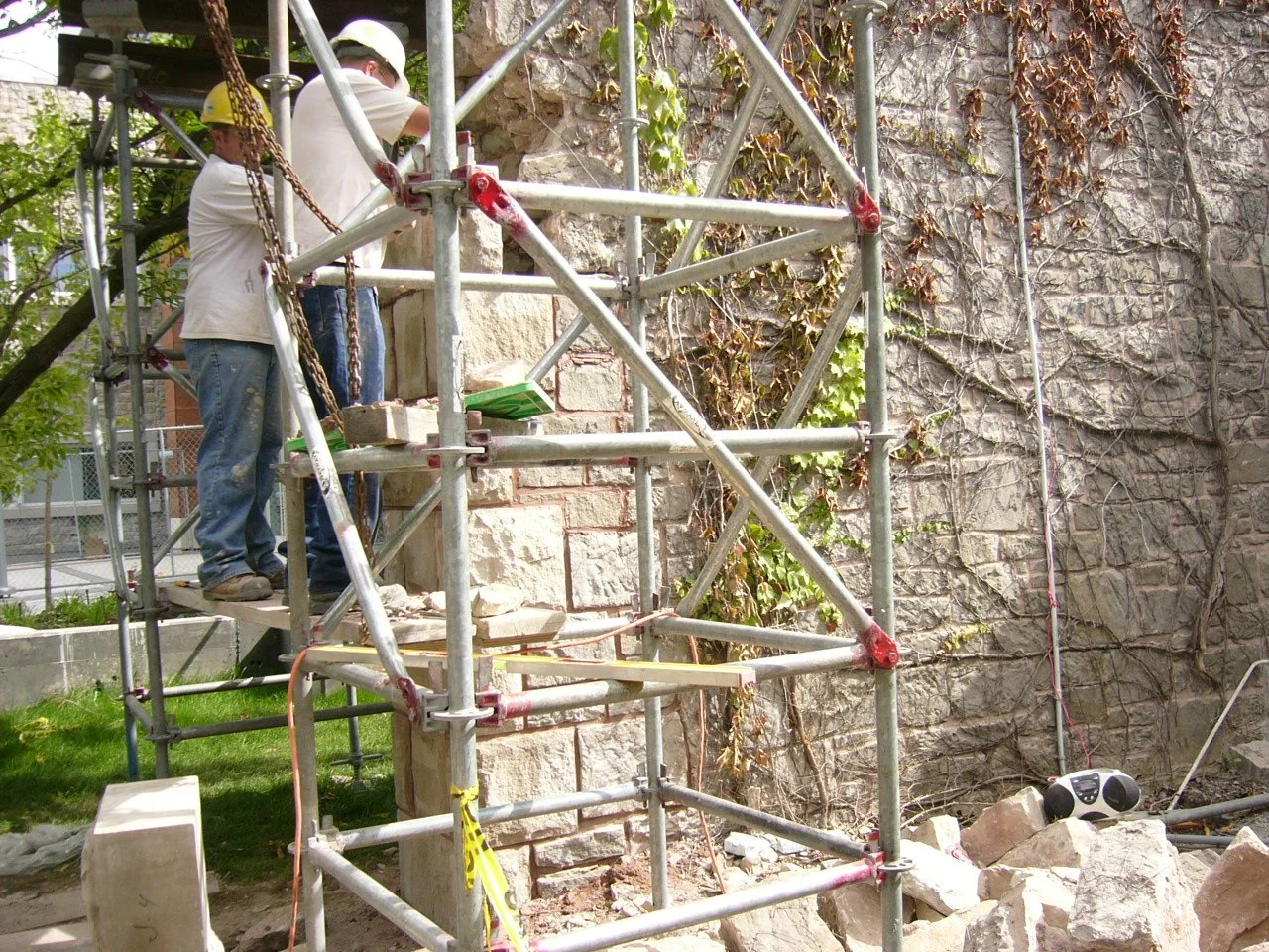 Two workers wearing hard hats on a scaffold working on a stone wall.