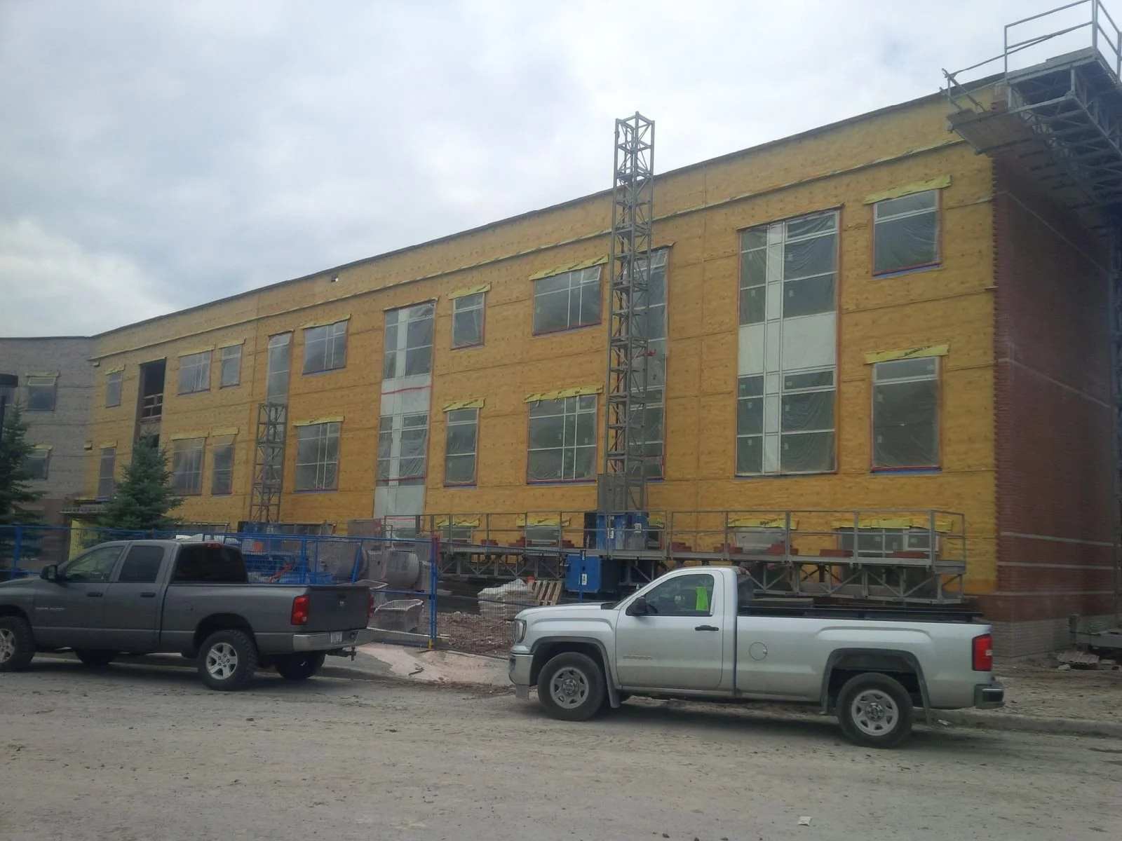Construction site with a building under renovation, yellow insulation on the exterior walls, large windows, two parked pickup trucks in the foreground, cloudy sky.