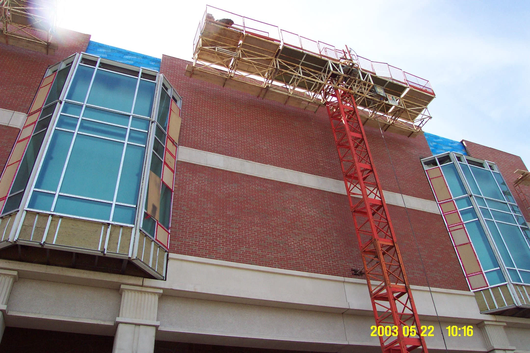 A building under construction with a large red tower crane, brick walls, and blue-tinted glass windows framed with red and beige panels.