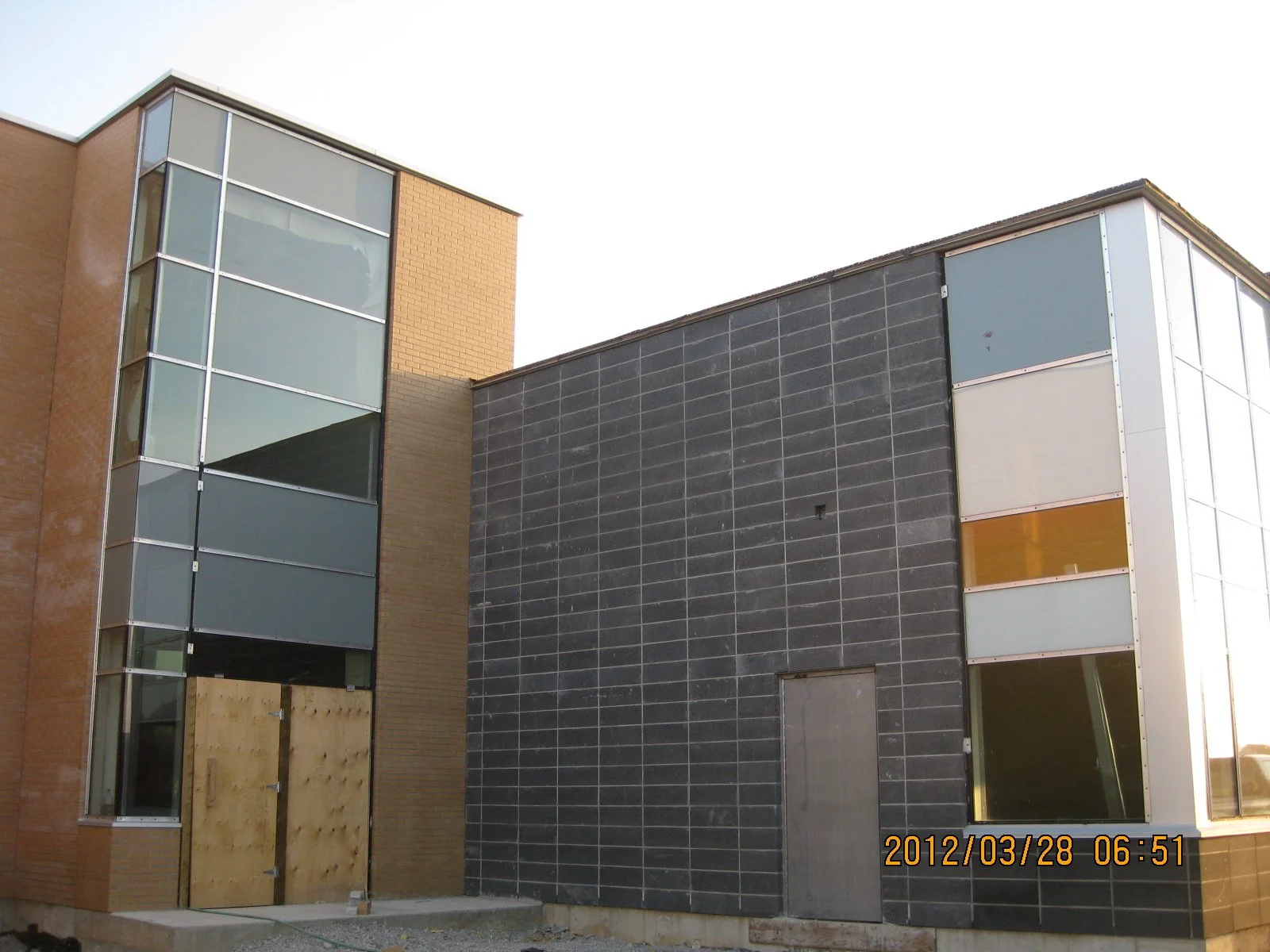Exterior of a modern building with large glass windows, brick, and dark gray tiled sections, and boarded-up door