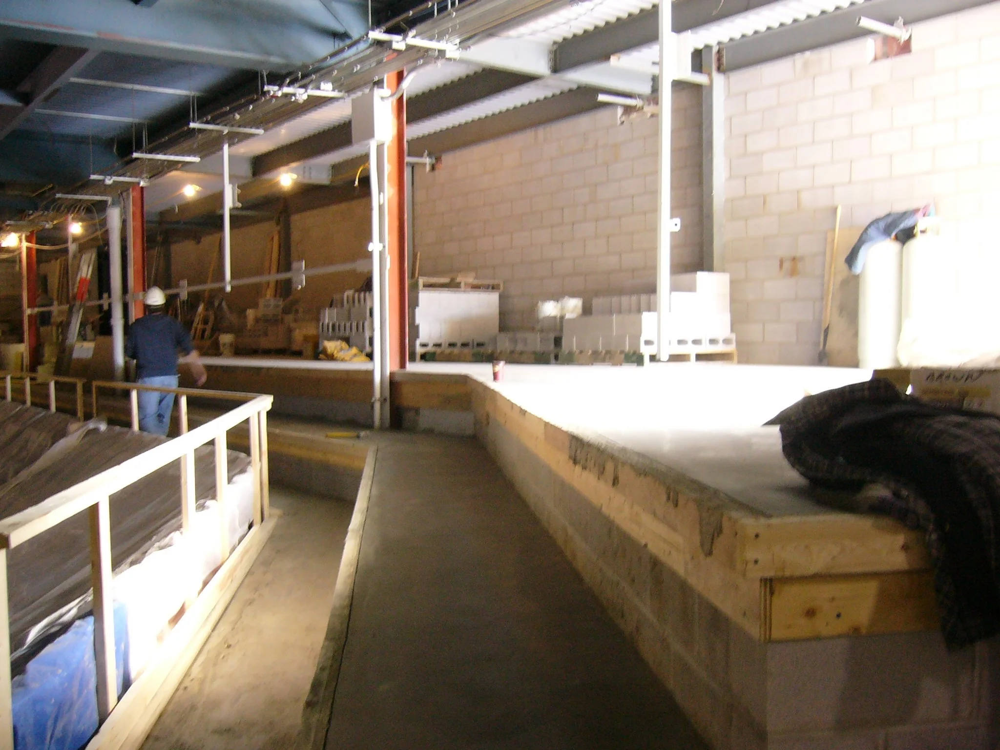 Construction site interior with a worker wearing a white helmet, unfinished walls, and building materials including cinder blocks and wooden beams.