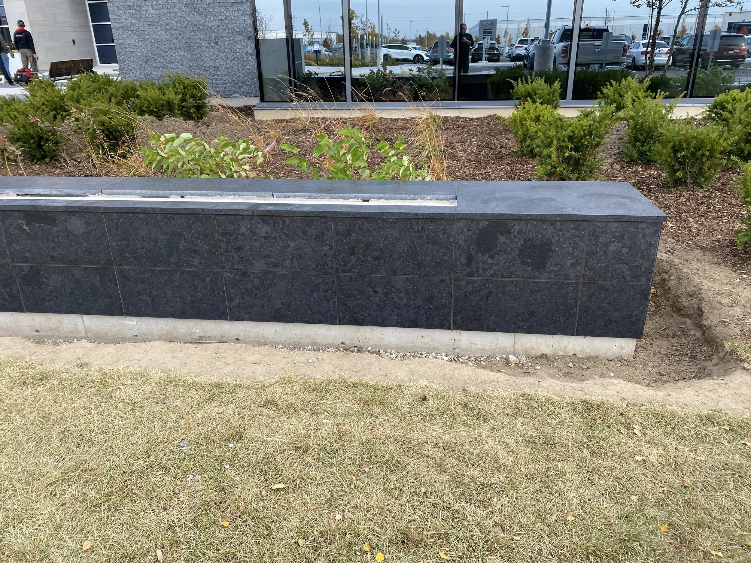 A rectangular black granite planter with plants inside, situated in front of a modern building with glass windows, a parking lot with cars, and a grassy area in the foreground.
