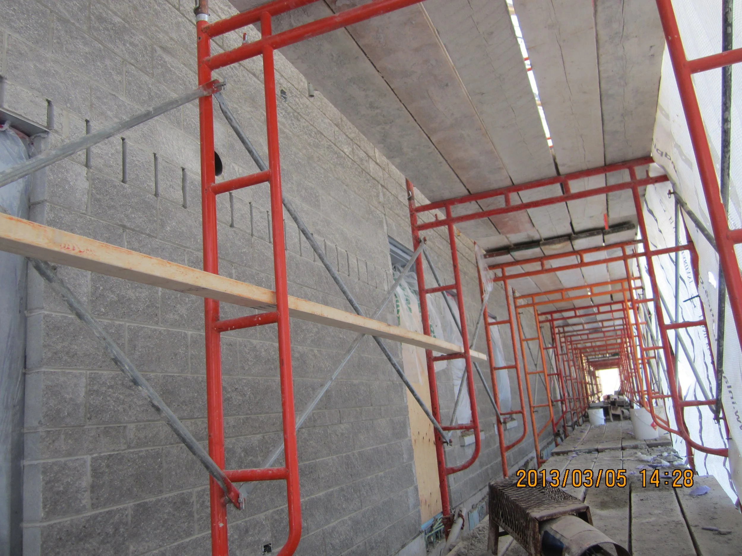 View of a construction site with orange scaffolding set up next to a brick wall, with construction tools and materials scattered on the ground.