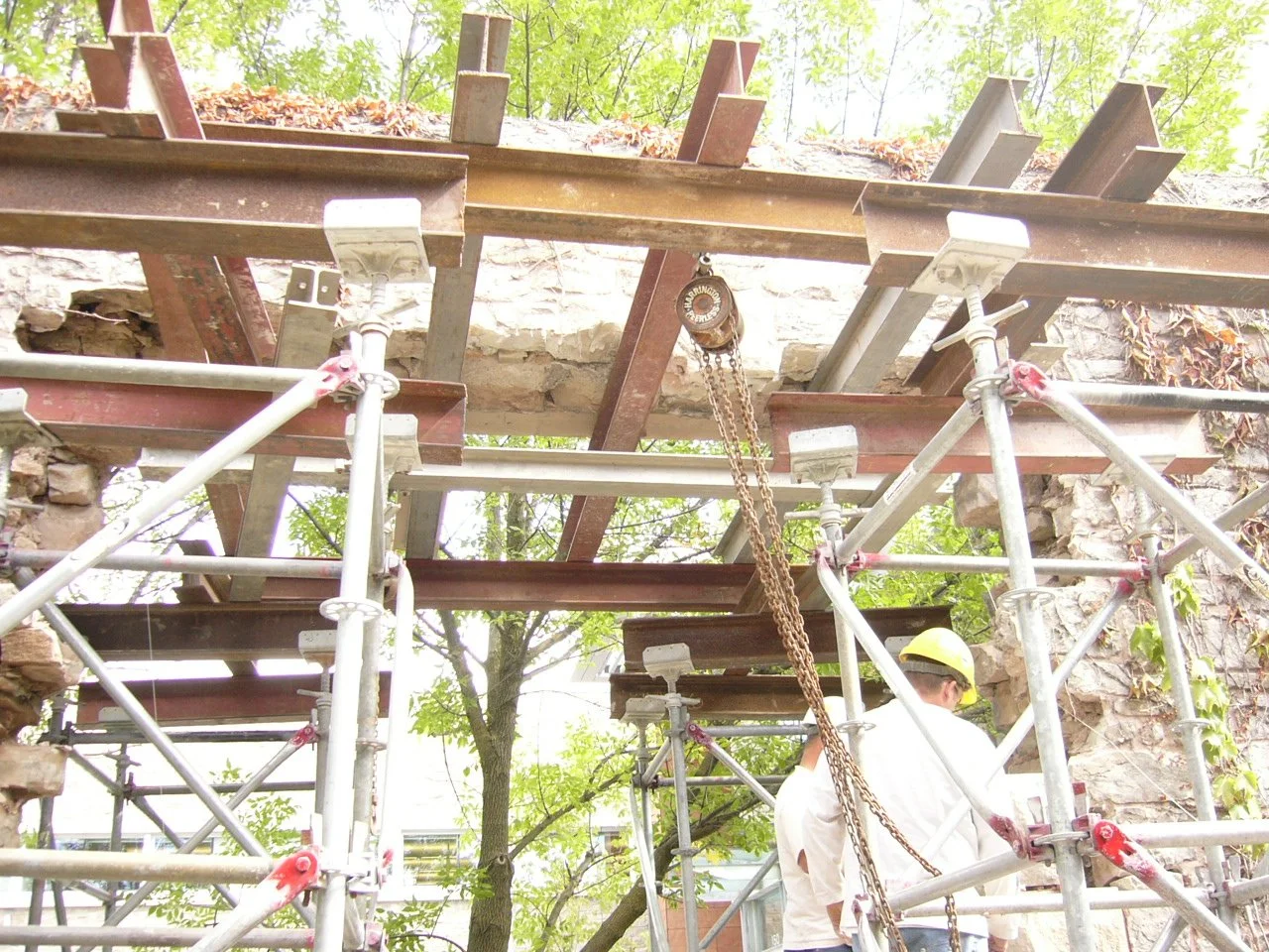 Construction workers inspecting the underside of an old stone and brick building supported by metal scaffolding and wooden beams.