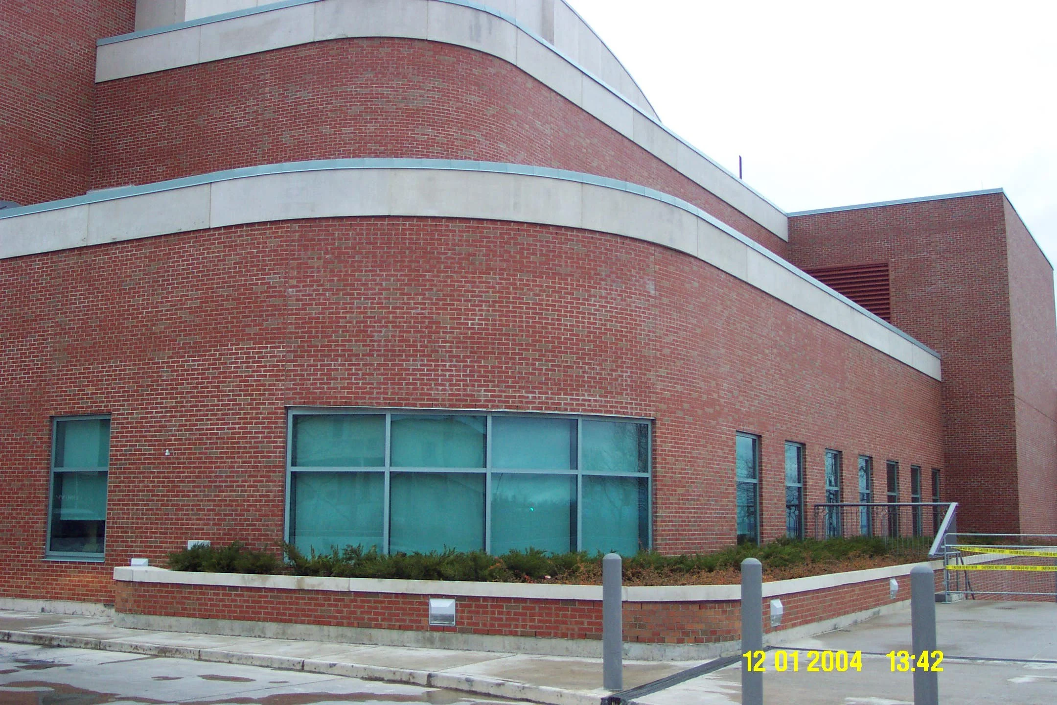Photo of a modern brick building with curved architecture, large windows, and concrete accents. The image includes a parking area with barriers and bollards, and a timestamp indicating it was taken on December 1, 2004, at 1:42 PM.