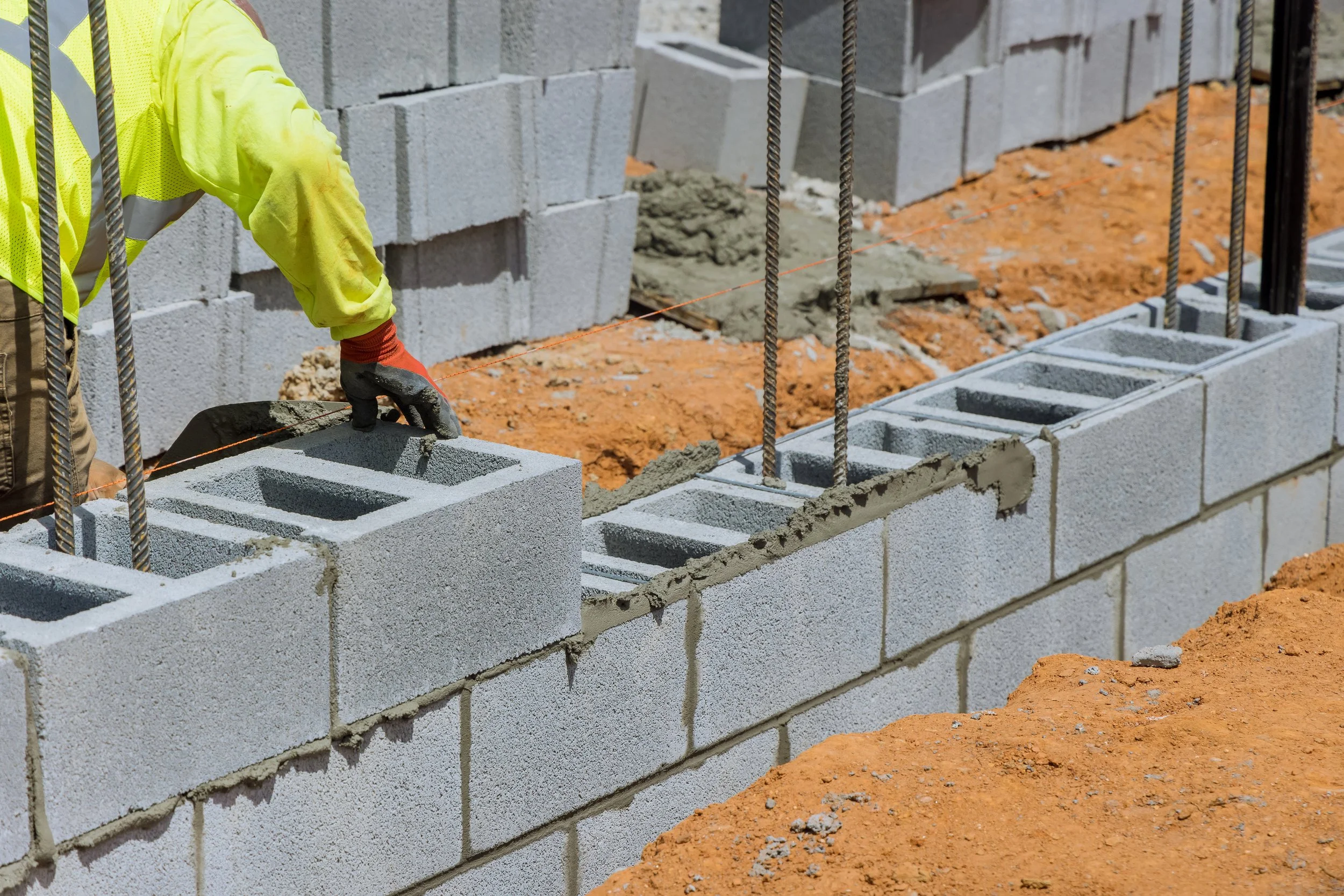Construction worker in a yellow safety vest and gloves building a concrete block wall, placing blocks and aligning them with a string line for level.