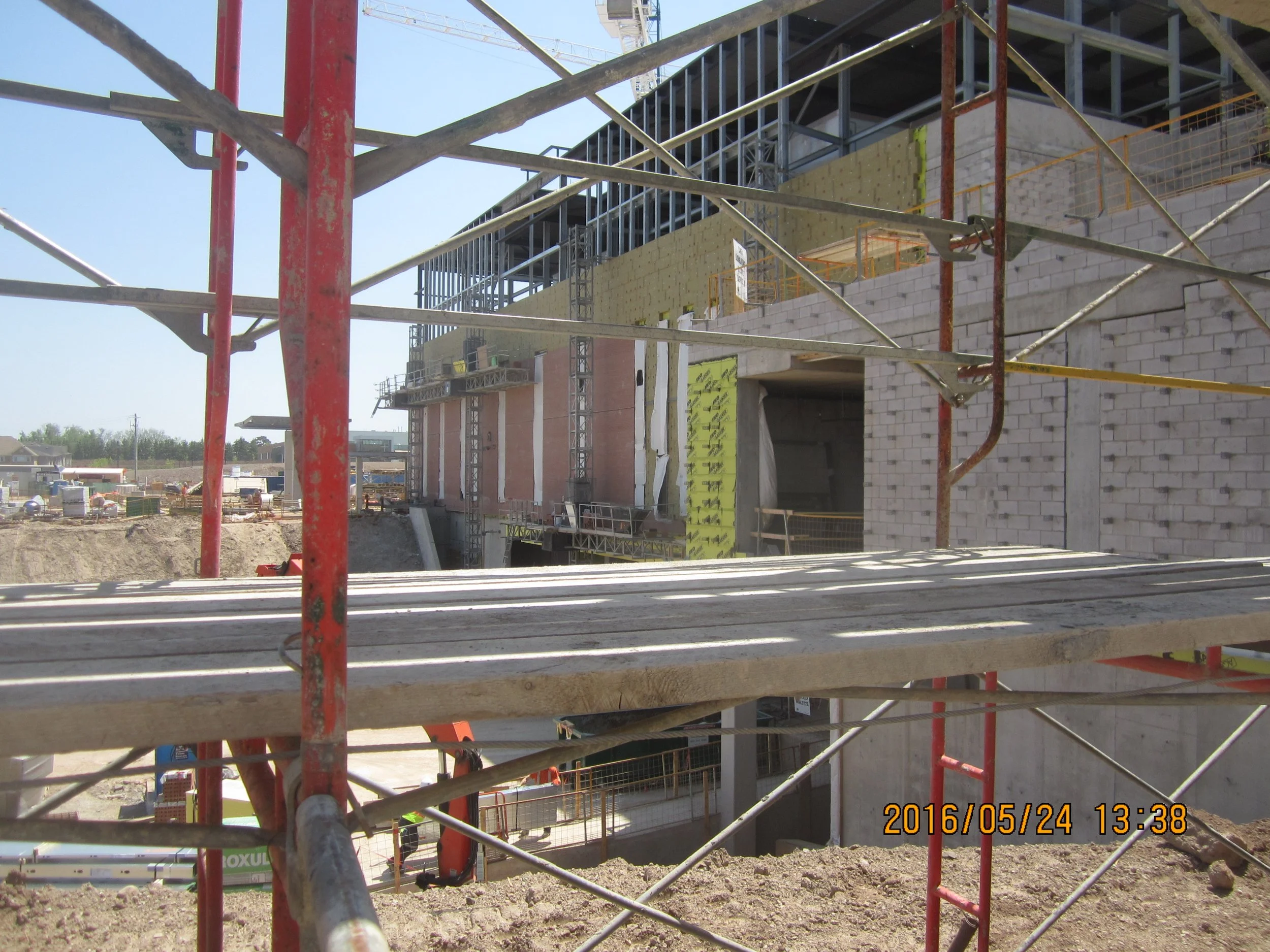 Construction site with scaffolding and partially built multi-story building, dirt ground, construction materials, and blue sky.