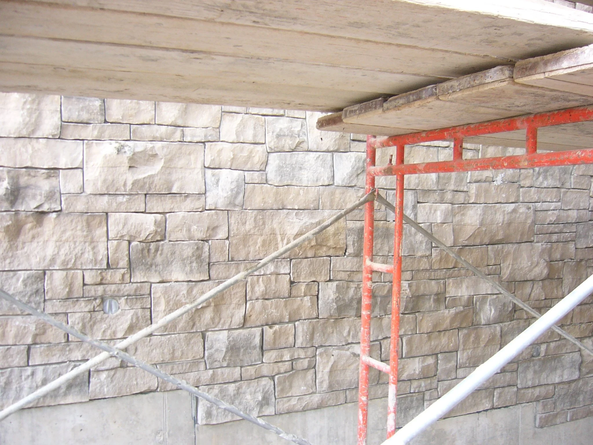A construction scaffold with red metal pipes and wooden planks in front of a stone wall.