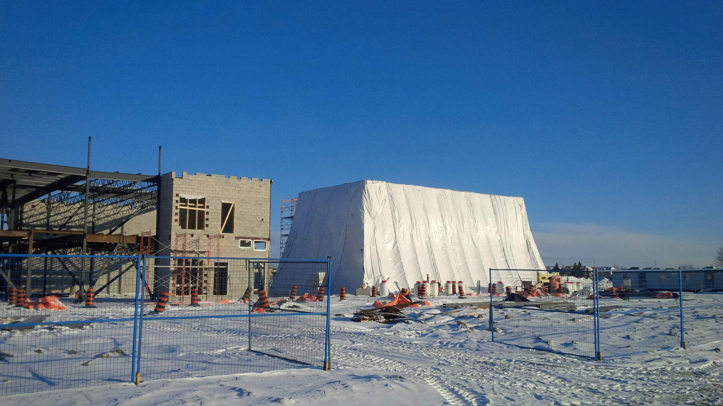 Construction site in winter with building under construction, scaffolding, and a large white tarp-covered structure surrounded by blue fencing and orange safety cones, snow on the ground, clear blue sky in the background.