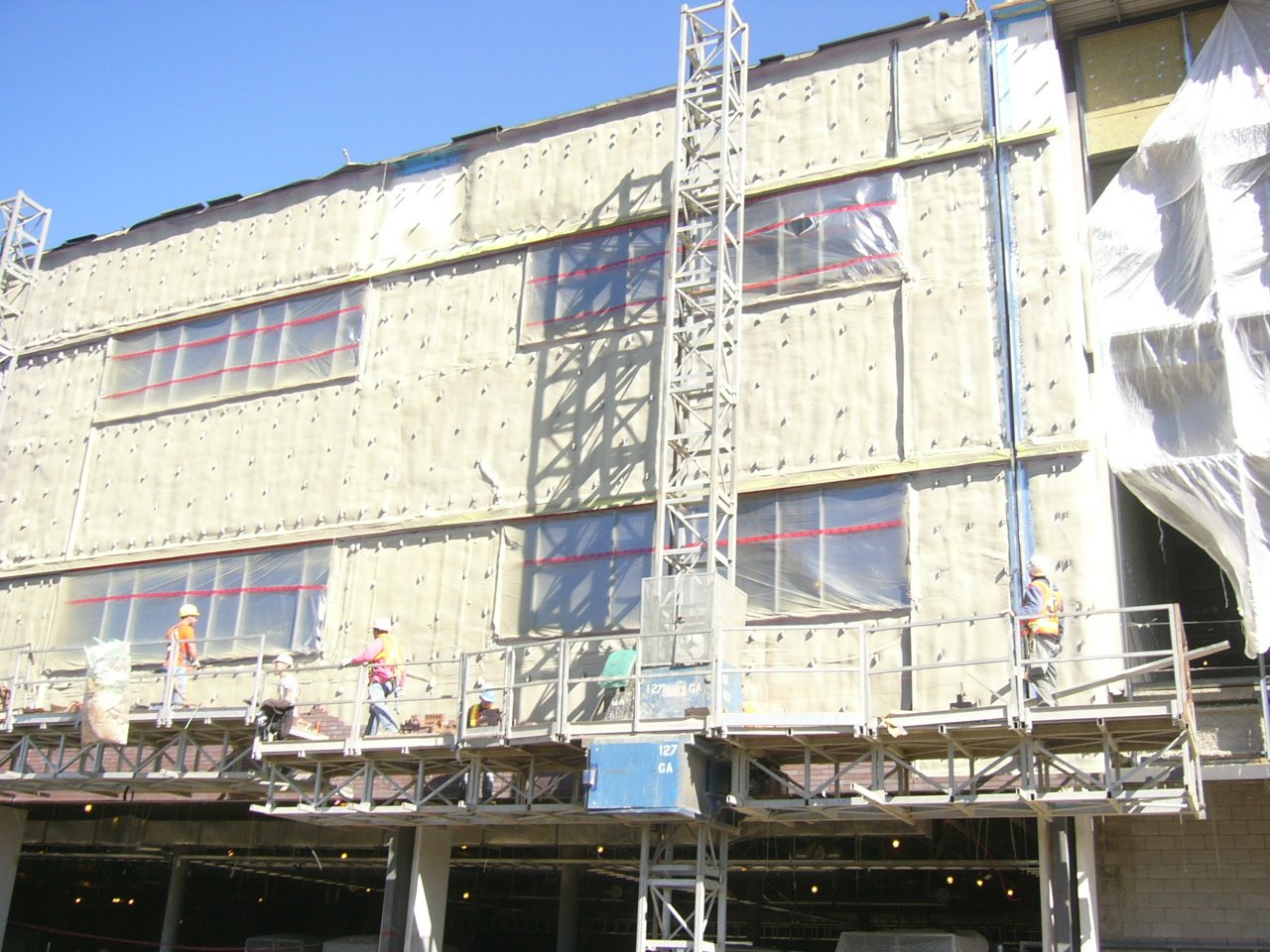 Construction workers wearing safety gear on scaffolding in front of a building under renovation, with wrapped windows and white sheeting on the right side, against a blue sky.