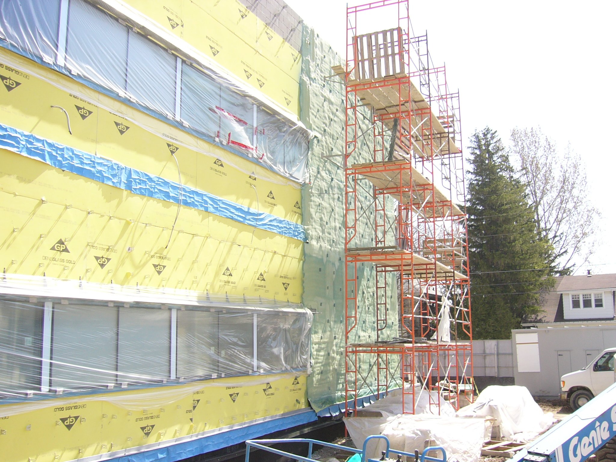 Construction site showing a building under renovations with yellow insulation and a red scaffolding tower next to it, with trees and houses in the background.