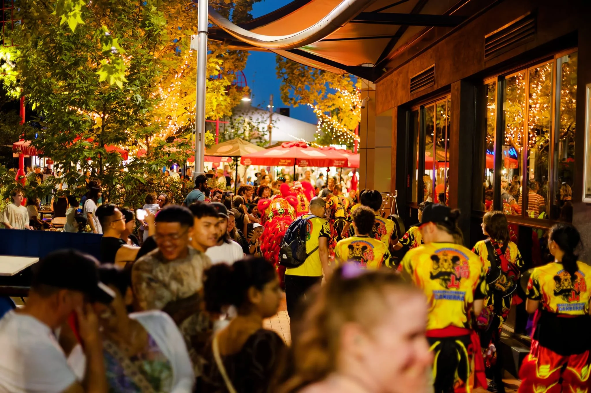 A crowded outdoor festival or celebration in the evening with people wearing colorful traditional costumes, some in yellow shirts with dragon motifs, gathering under string lights and red umbrellas, with trees and a building nearby.
