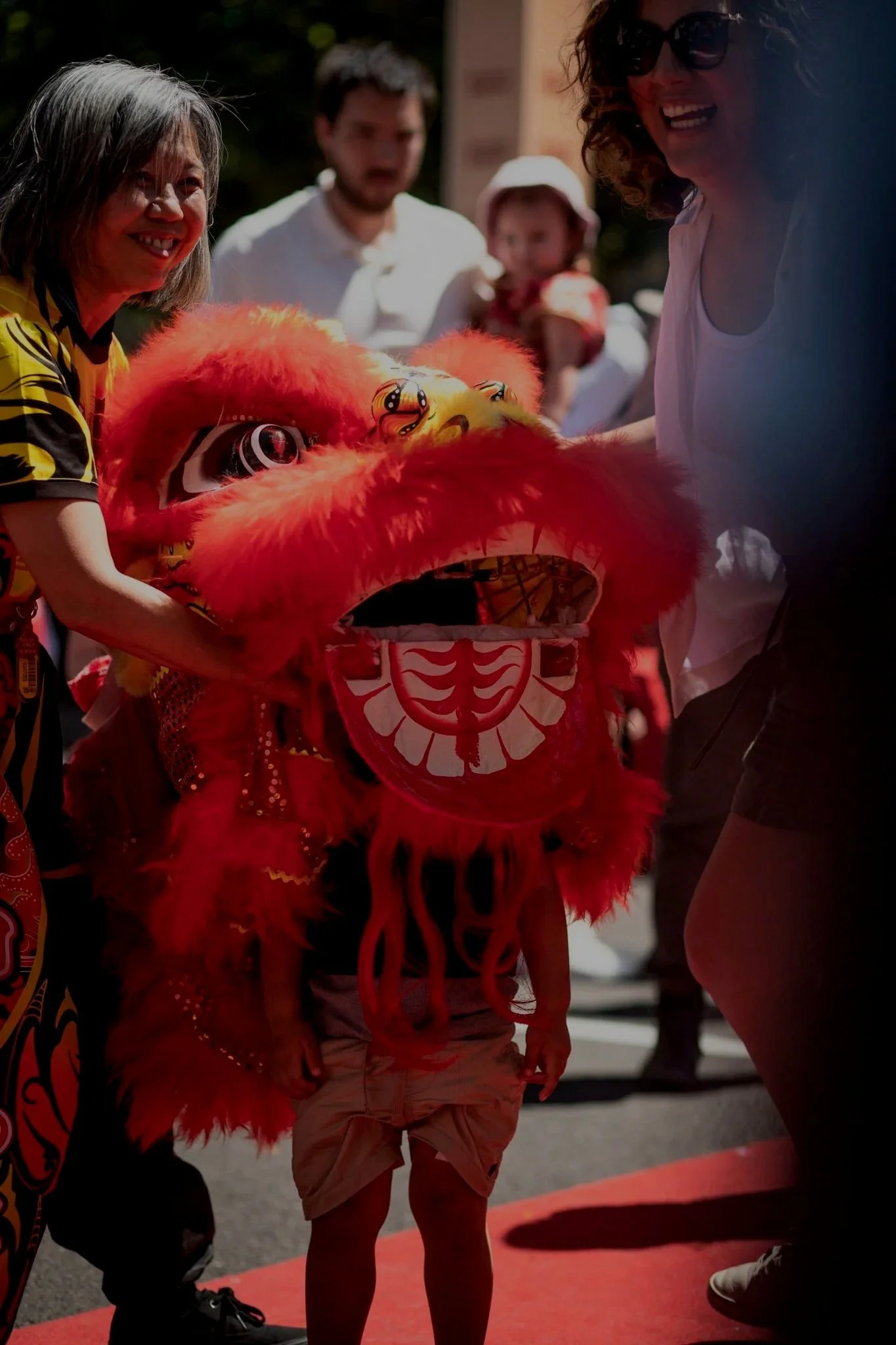 A young child participating in a traditional Chinese lion dance. The child is standing under a red lion costume with a large, colorful head, while two women hold the lion's head for the performance. Other people are visible in the background, watching and smiling during a festive outdoor event.