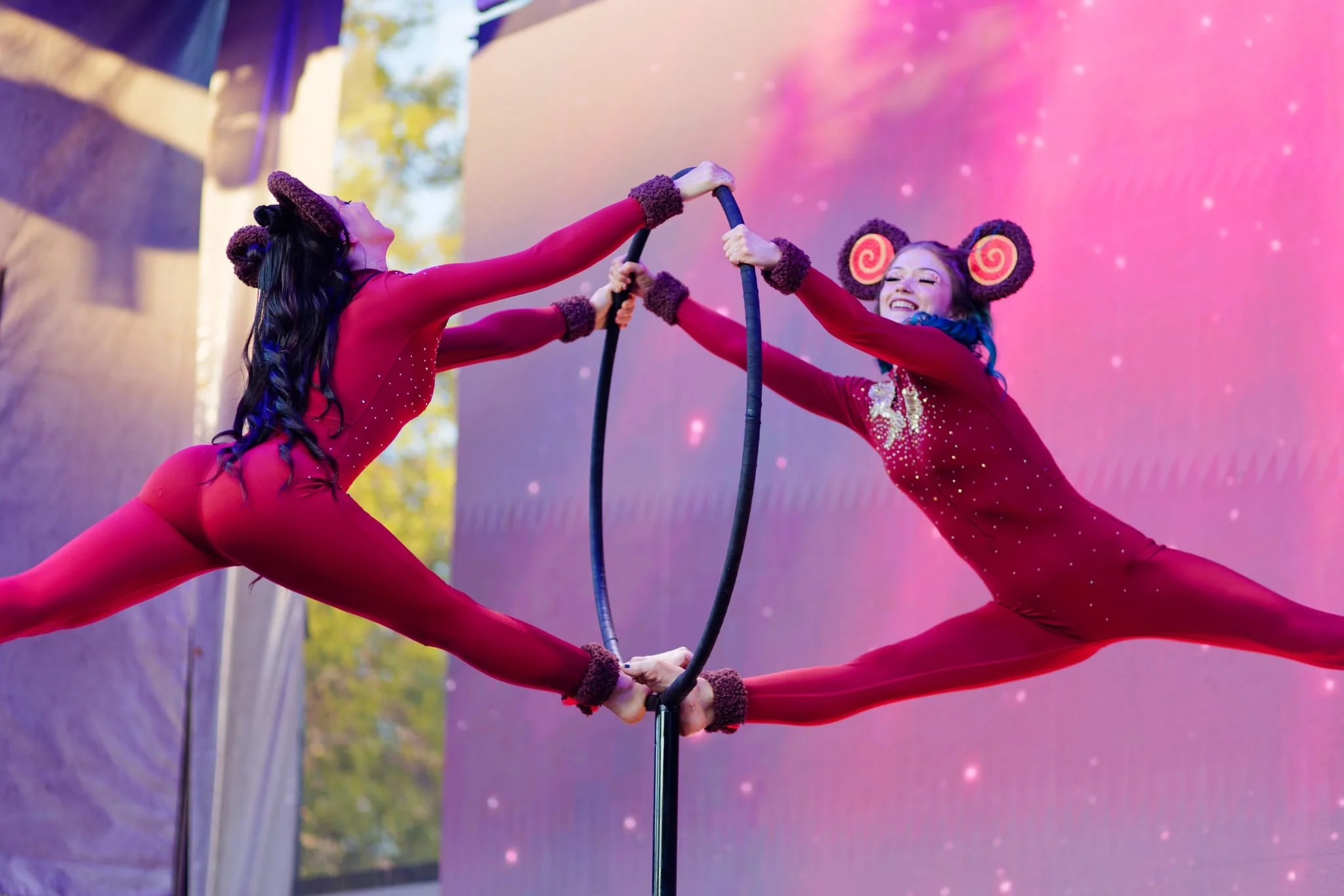 Two female acrobats performing on a Cyr wheel on stage, wearing matching red costumes with fairy tale-like headgear, against a pink and purple cosmic background.