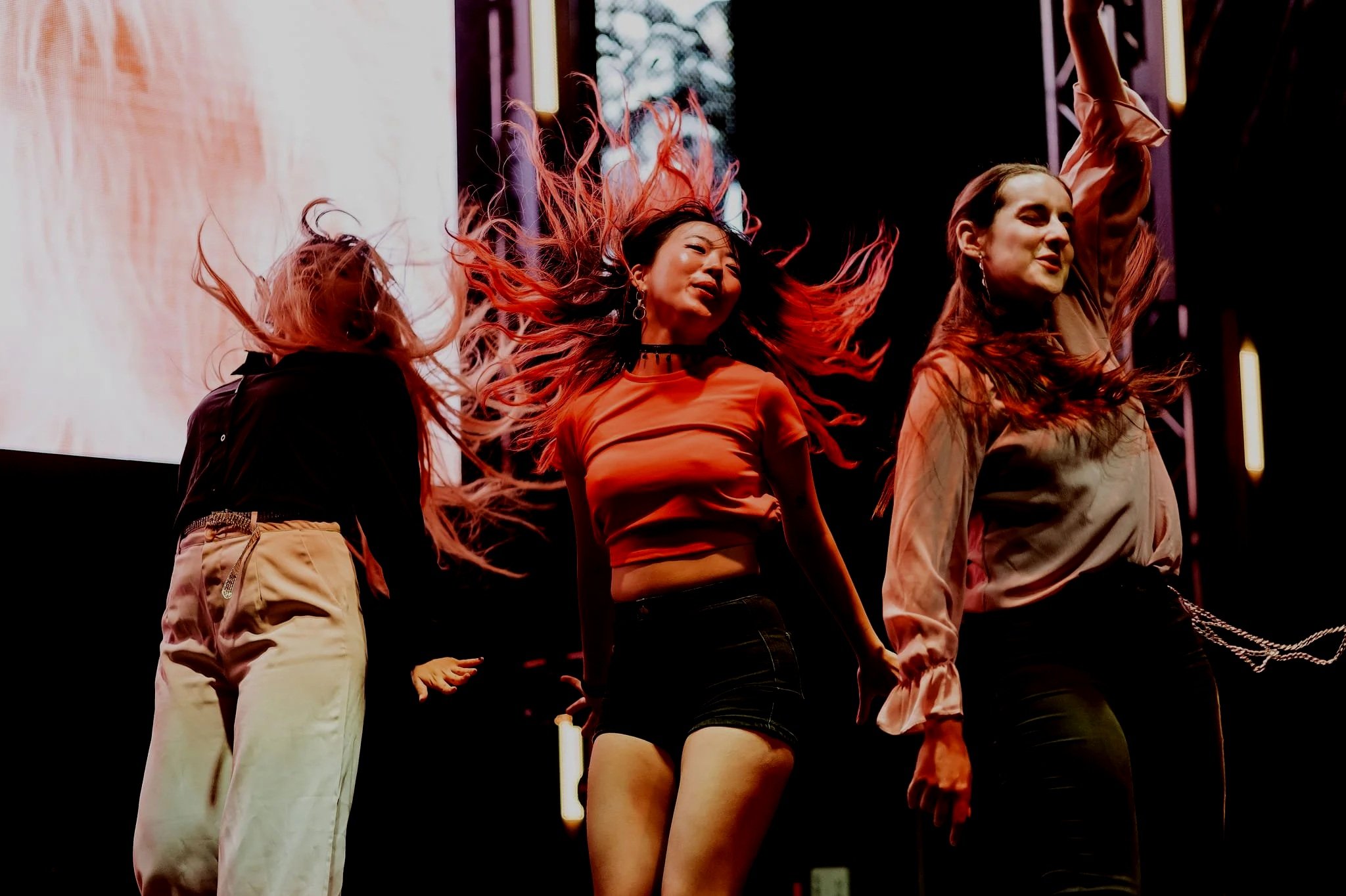 Three women dancing on stage with vibrant hair movements, illuminated by stage lights, in a lively performance setting.