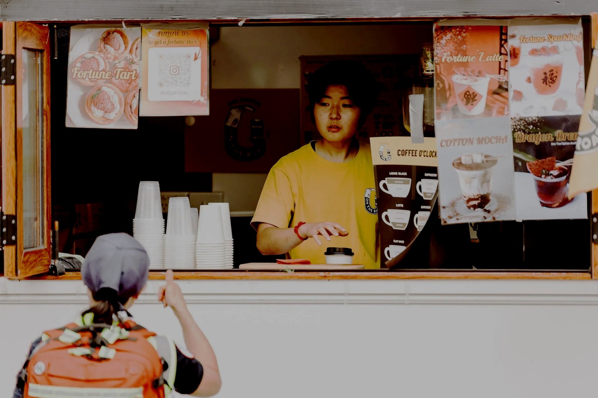A young girl in a cap and backpack points at a food truck window where a female worker in a yellow shirt is preparing a beverage. The food truck has various posters of drinks including Fortune Latte, Cotton Mocha, and Dragon Brew.