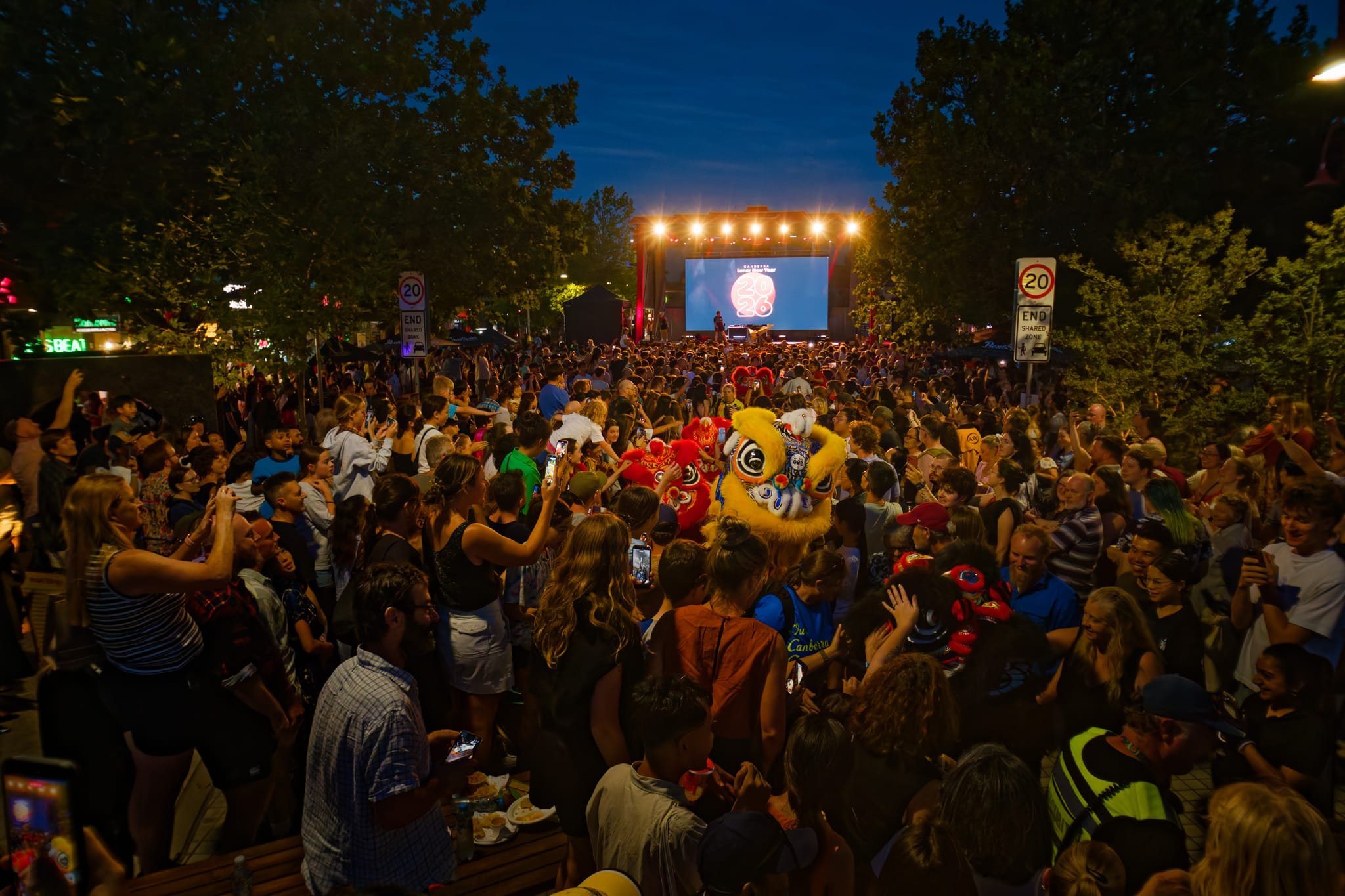 Crowd of people gathered outdoors at night watching stage with a lion dance performance, some using phones to take photos or videos, trees and signs surrounding the scene.