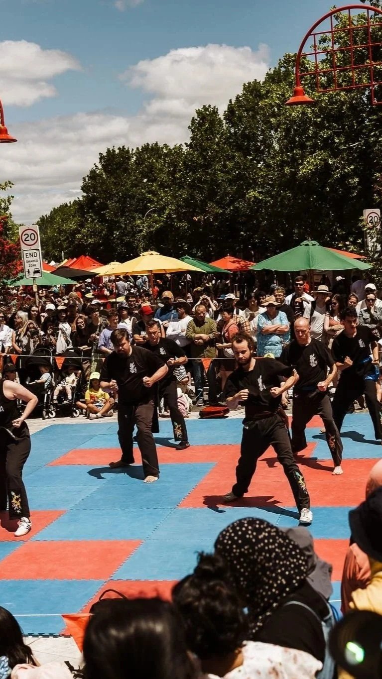A martial arts demonstration or performance taking place outdoors on a sunny day, with several practitioners in black uniforms performing in front of a large crowd under colorful umbrellas and trees.