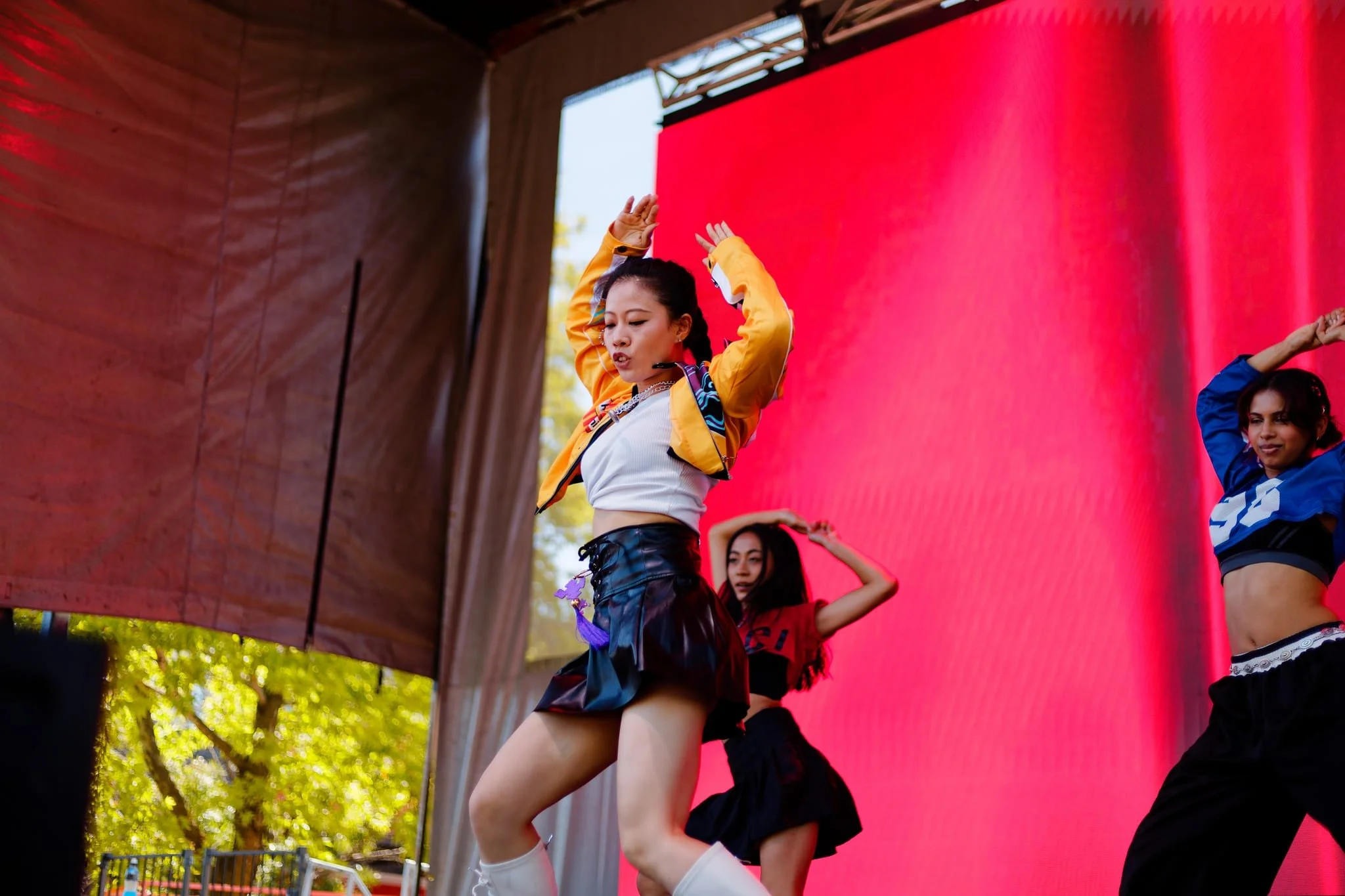 Group of young women performing a dance on stage outdoors with a large red screen in the background.