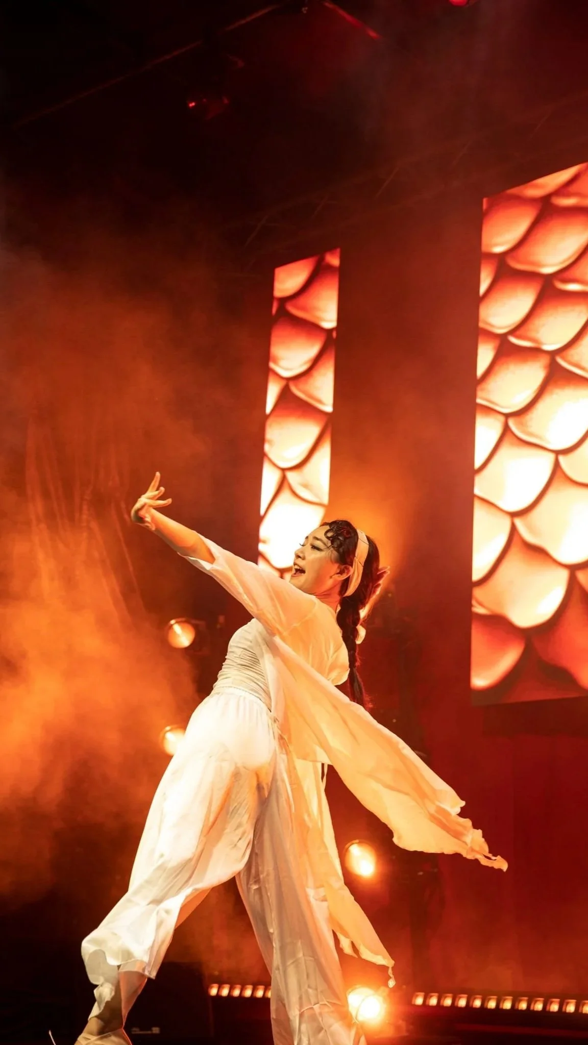 Performer in flowing white costume dancing on stage with orange lighting and large abstract electronic screens in the background.