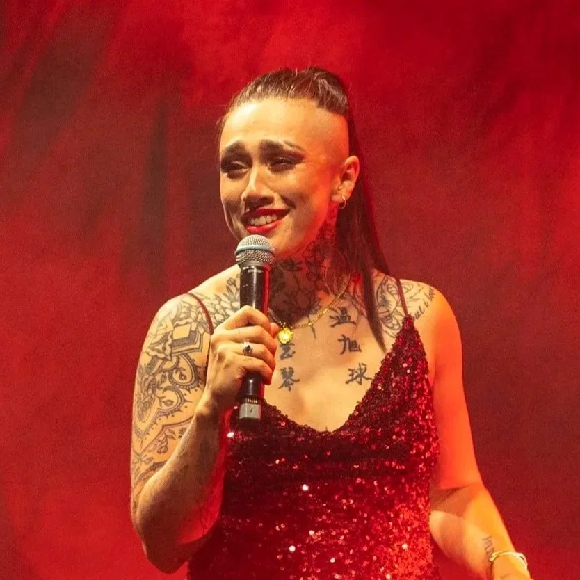 A person  wearing a red sequin dress, holding a microphone, and smiling on stage with red lighting.