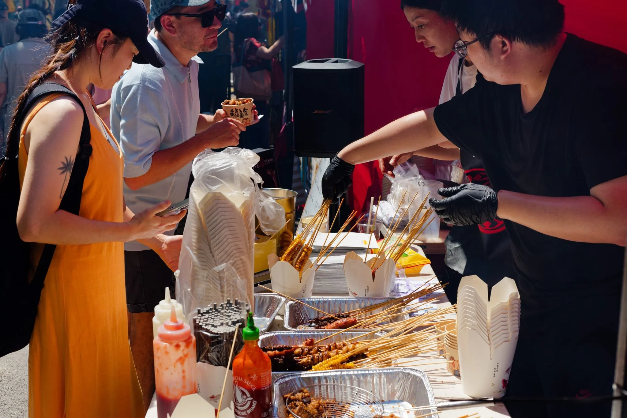 People at a street food stall selecting skewered grilled meat and snacks, with condiments and utensils on the table.