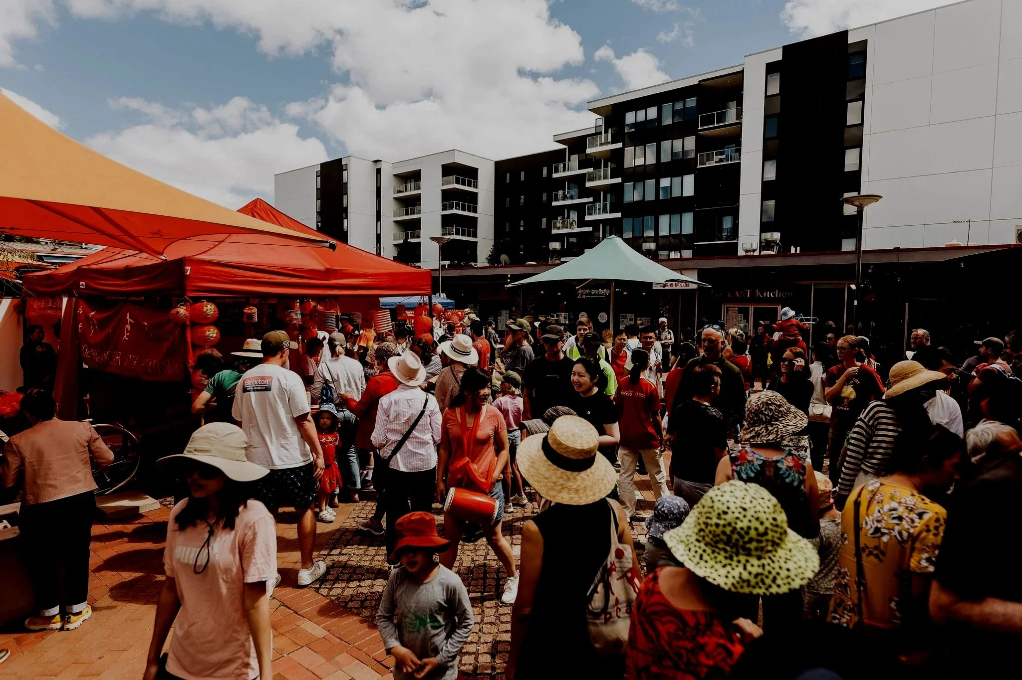 Crowd of people at an outdoor market or festival with red and white tents and modern apartment buildings in the background.