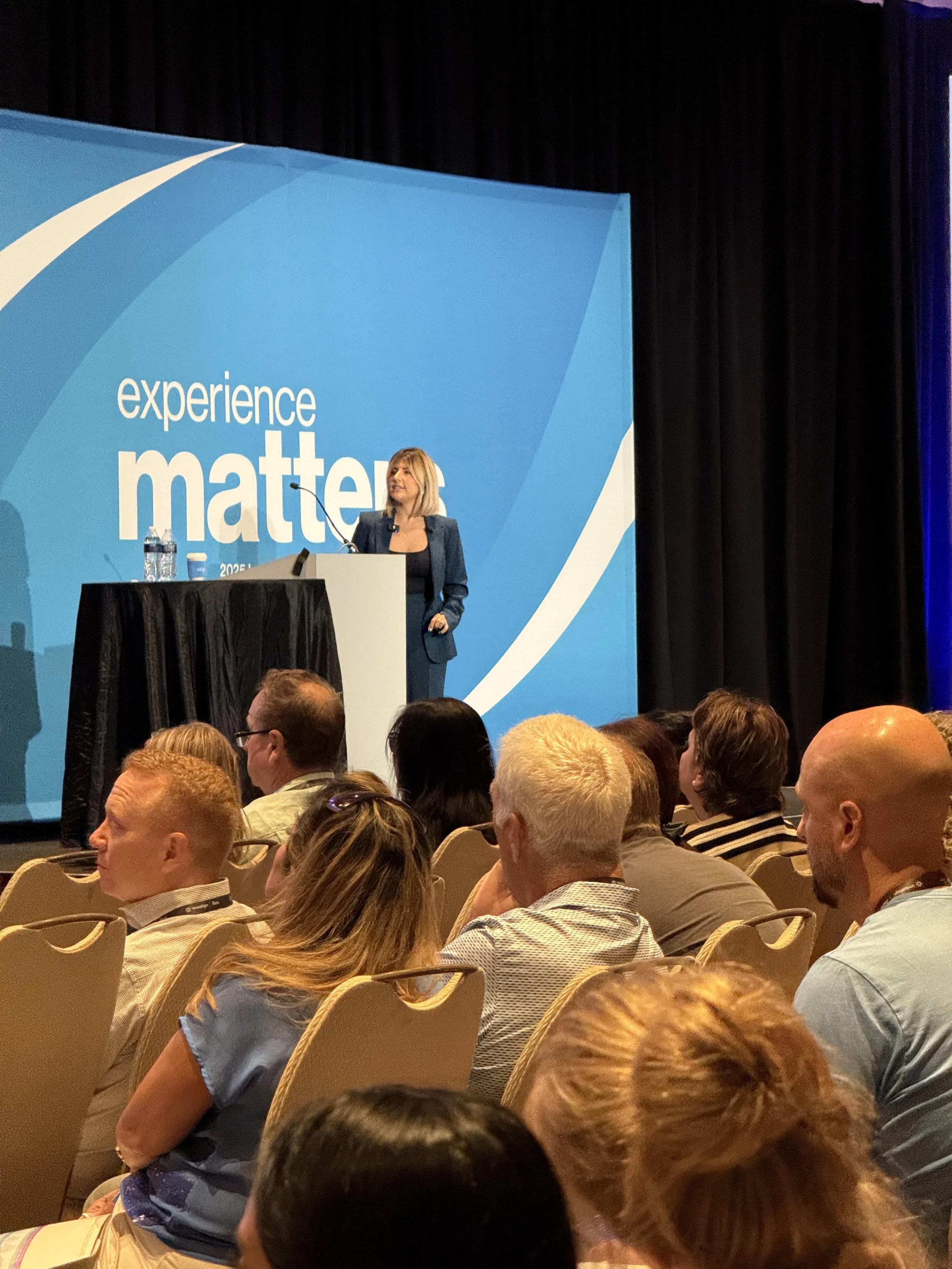A woman giving a speech or presentation at a conference, standing at a podium in front of a large blue backdrop with the words 'experience matter'.