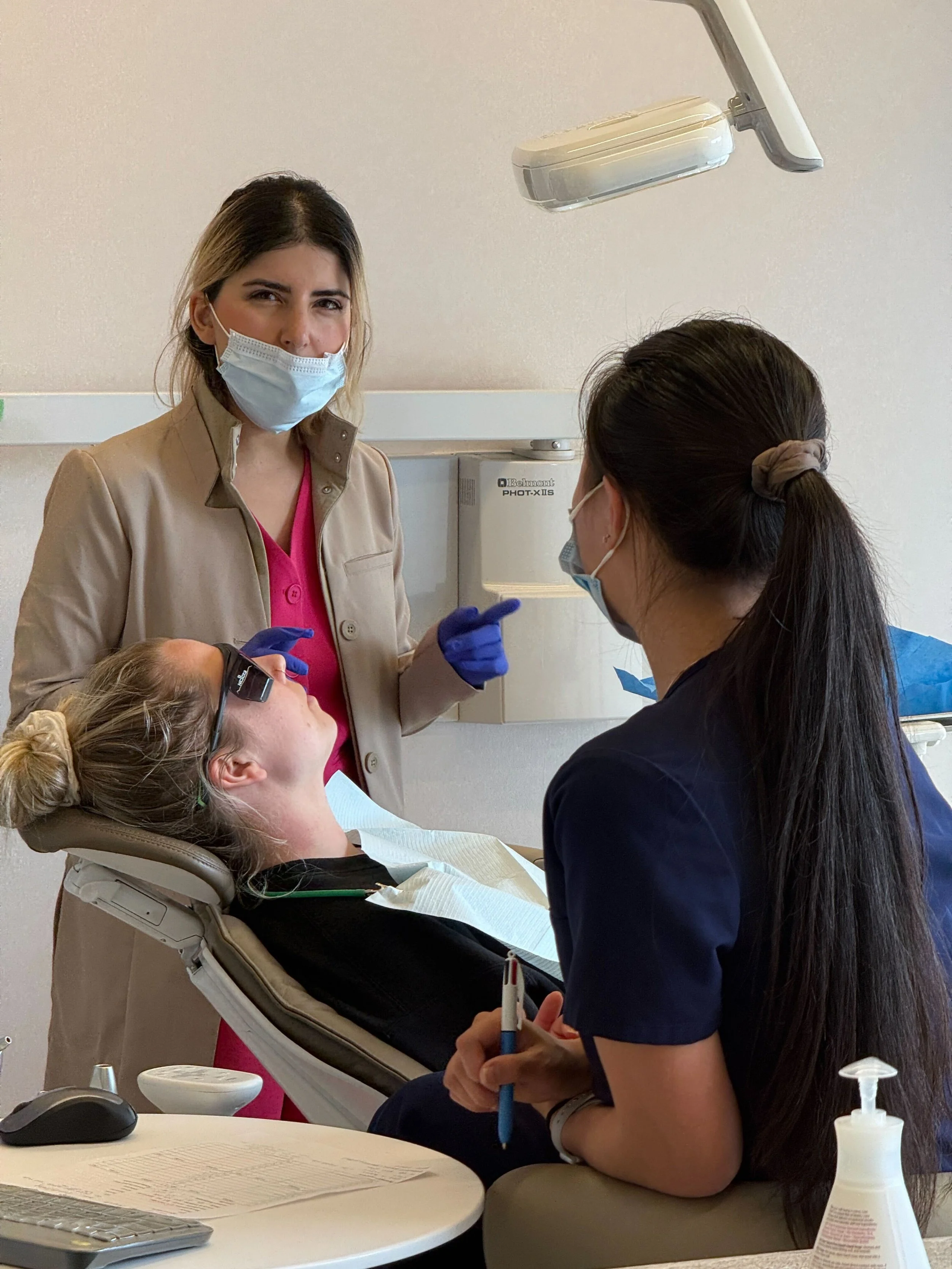 A woman lying back in a dental chair at a dental clinic, with her mouth open for dental treatment. A female dental professional standing beside her, wearing a face mask, glasses, and gloves, is preparing to examine or treat her. There are dental tools and supplies on a small table nearby.