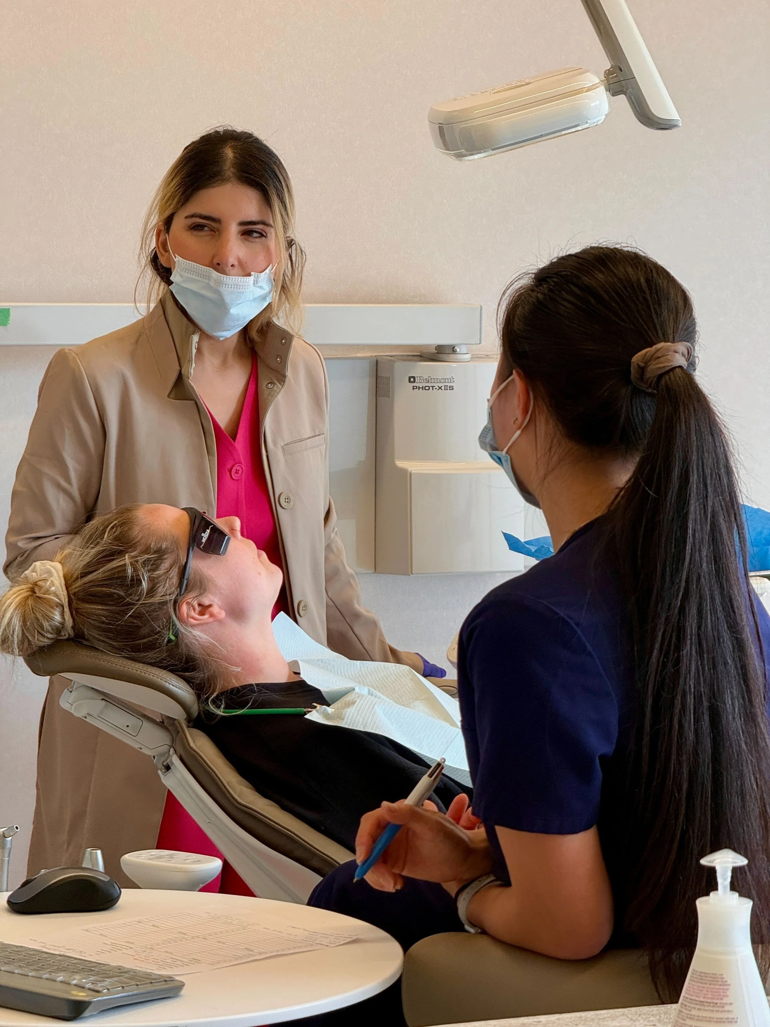 A woman lying in a dental chair with a dental bib, wearing protective glasses and a hair tie. A dental professional and another person wearing face masks are attending to her, with the professional holding dental tools and the other taking notes or managing equipment. There are dental and office supplies on a nearby table.
