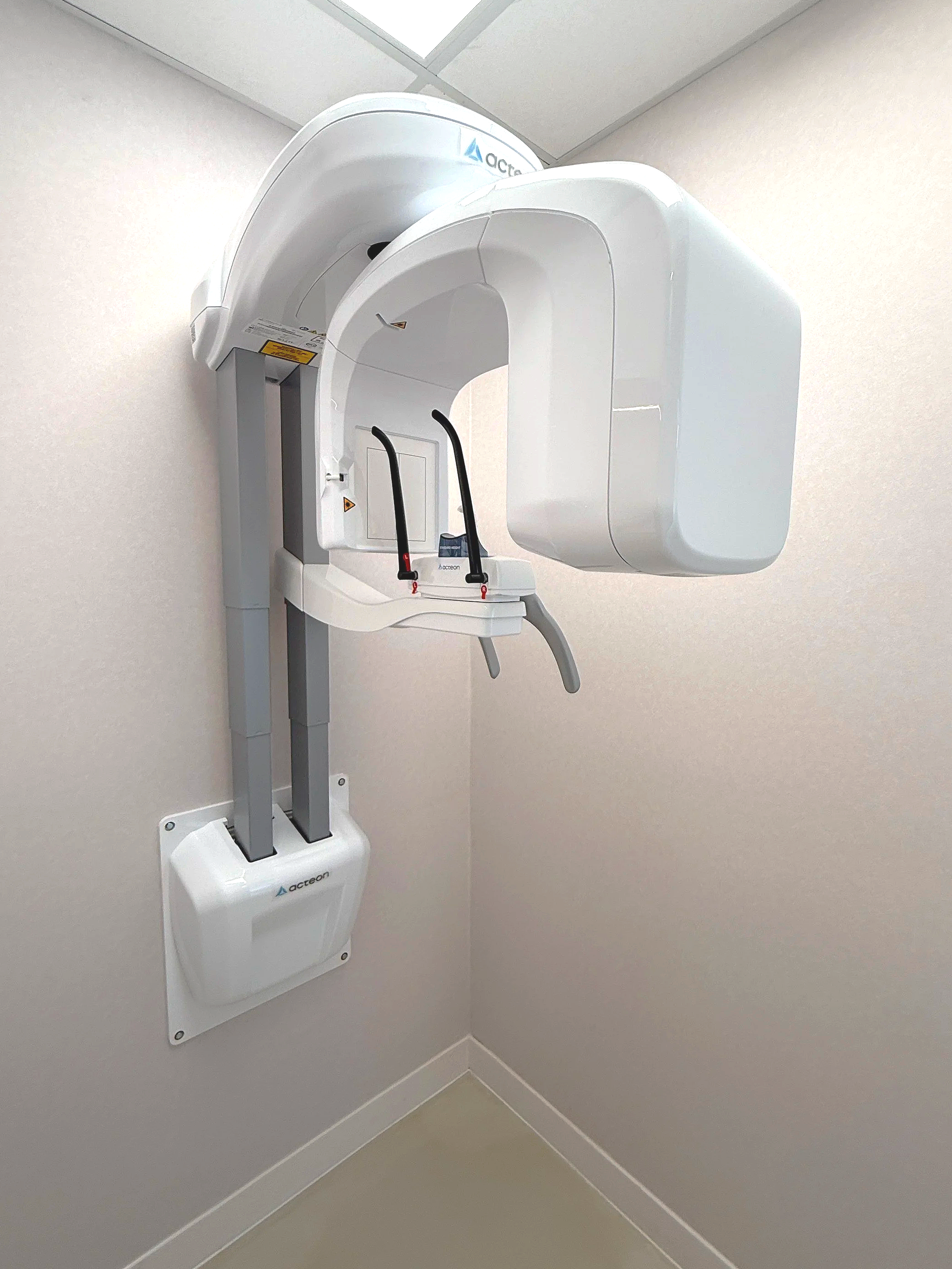 A panoramic dental X-ray machine mounted on the wall in a corner of a room with light-colored walls and a ceiling.