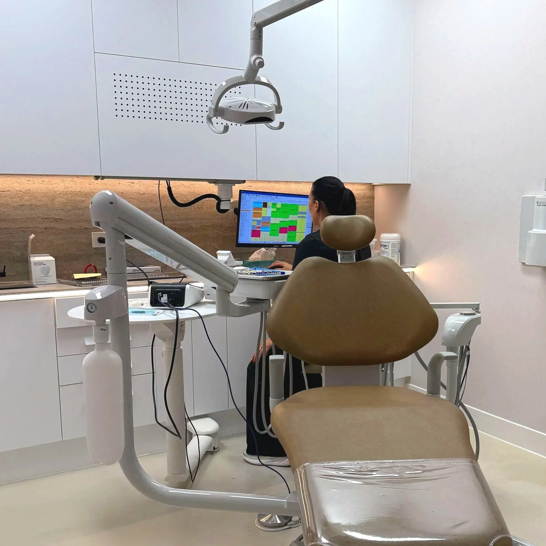 A dental examination room with a dental chair, overhead light, and a computer monitor showing dental records. A person, possibly a dentist or dental assistant, is working at the computer. The room has white cabinets and dental equipment.