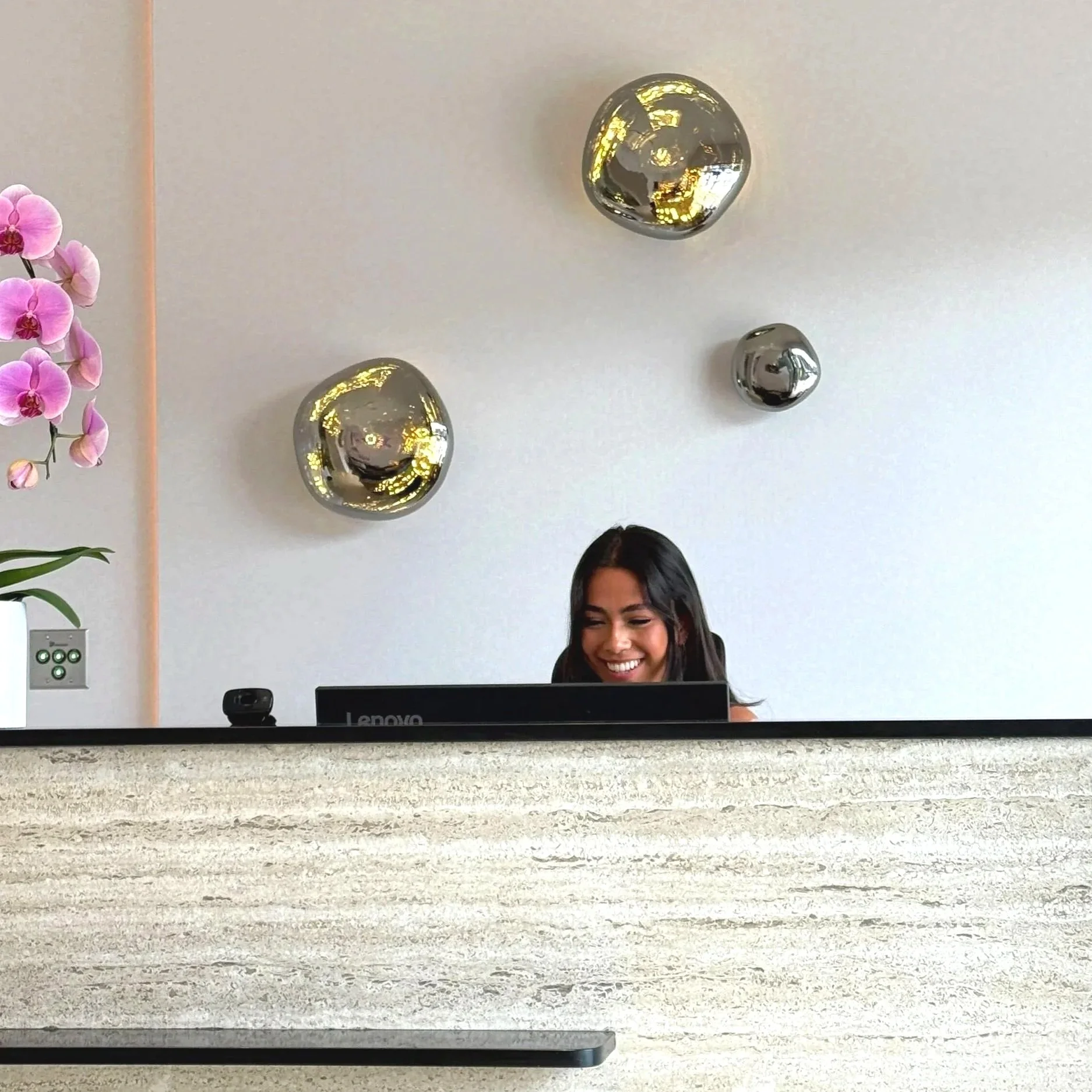 A woman with long dark hair smiling while working on a laptop at a desk with pink orchids, a black camera, and a computer monitor in a modern office with decorative silver spheres and a power outlet on the wall.
