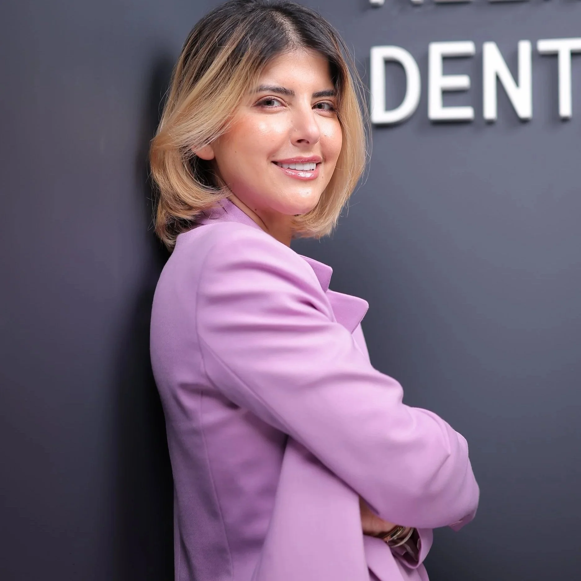 A woman wearing a pink blazer with blonde hair styled in a bob, smiling at the camera, standing against a black wall with white letters partially visible.