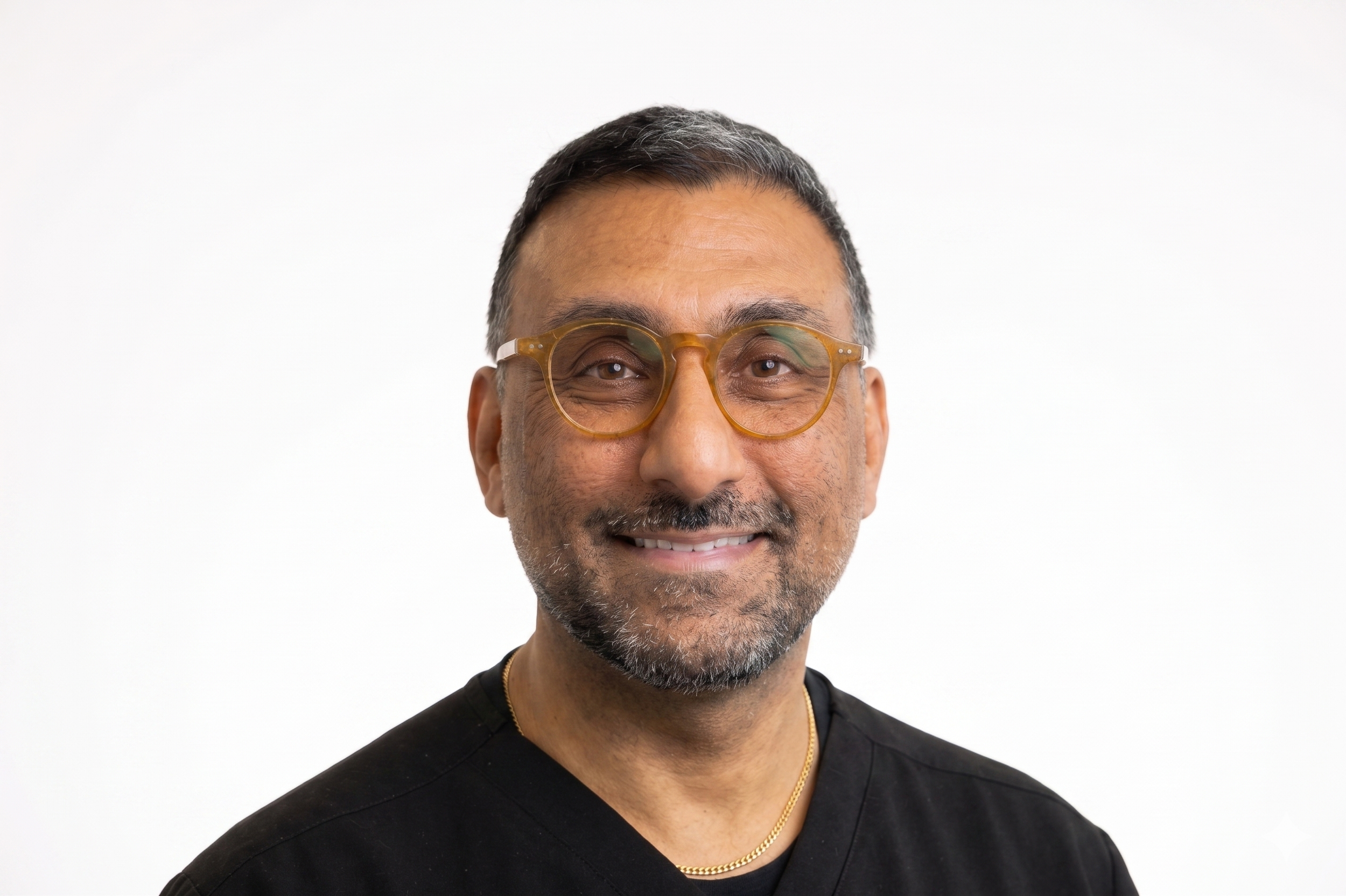 A smiling middle-aged man with salt-and-pepper hair, wearing glasses with yellow frames, a black shirt, and a gold chain necklace against a plain white background.