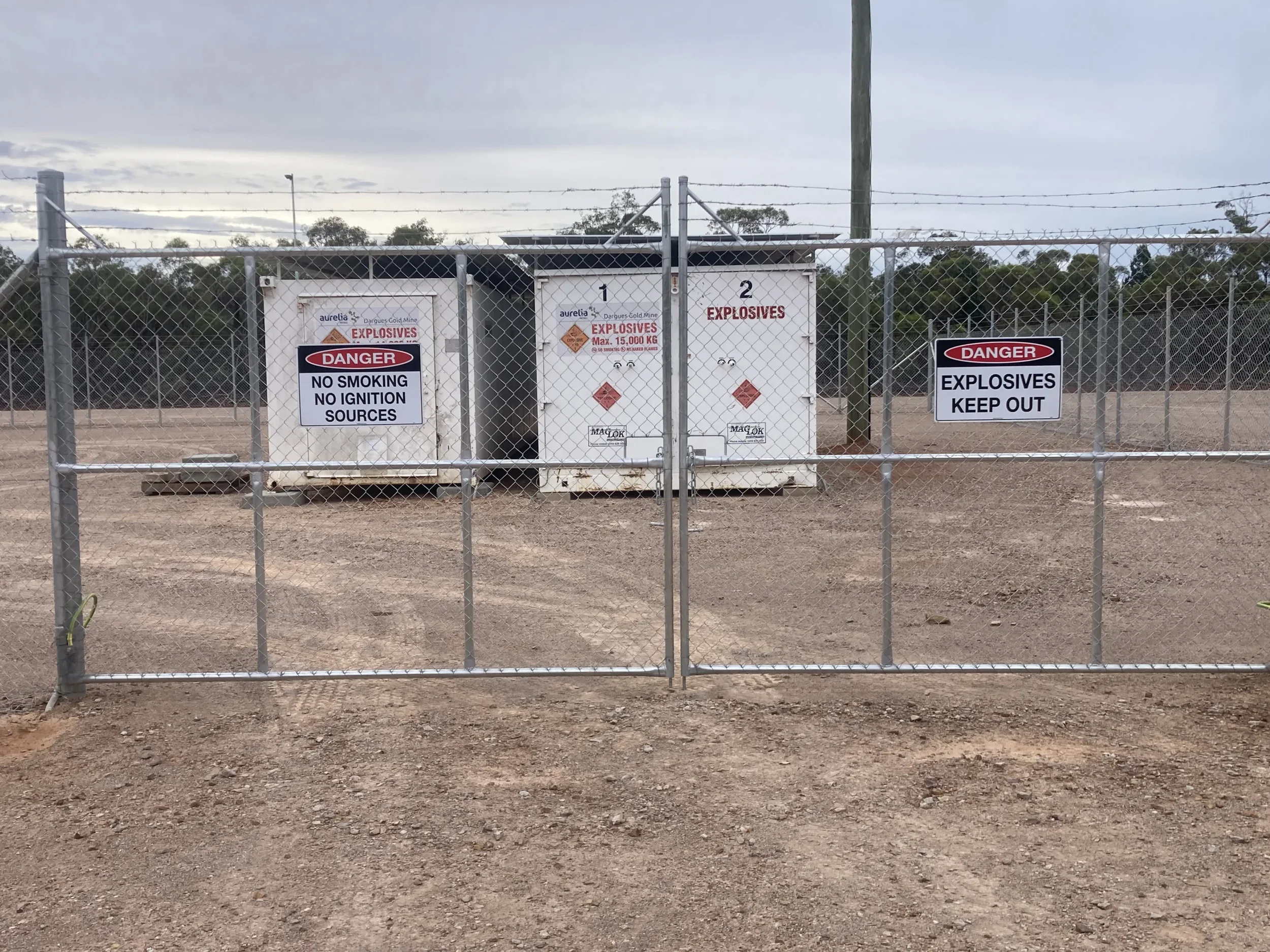 A metal security fence with signs warning about explosives, no smoking, and no ignition sources, enclosing a fenced area with trees and cloudy sky in the background.