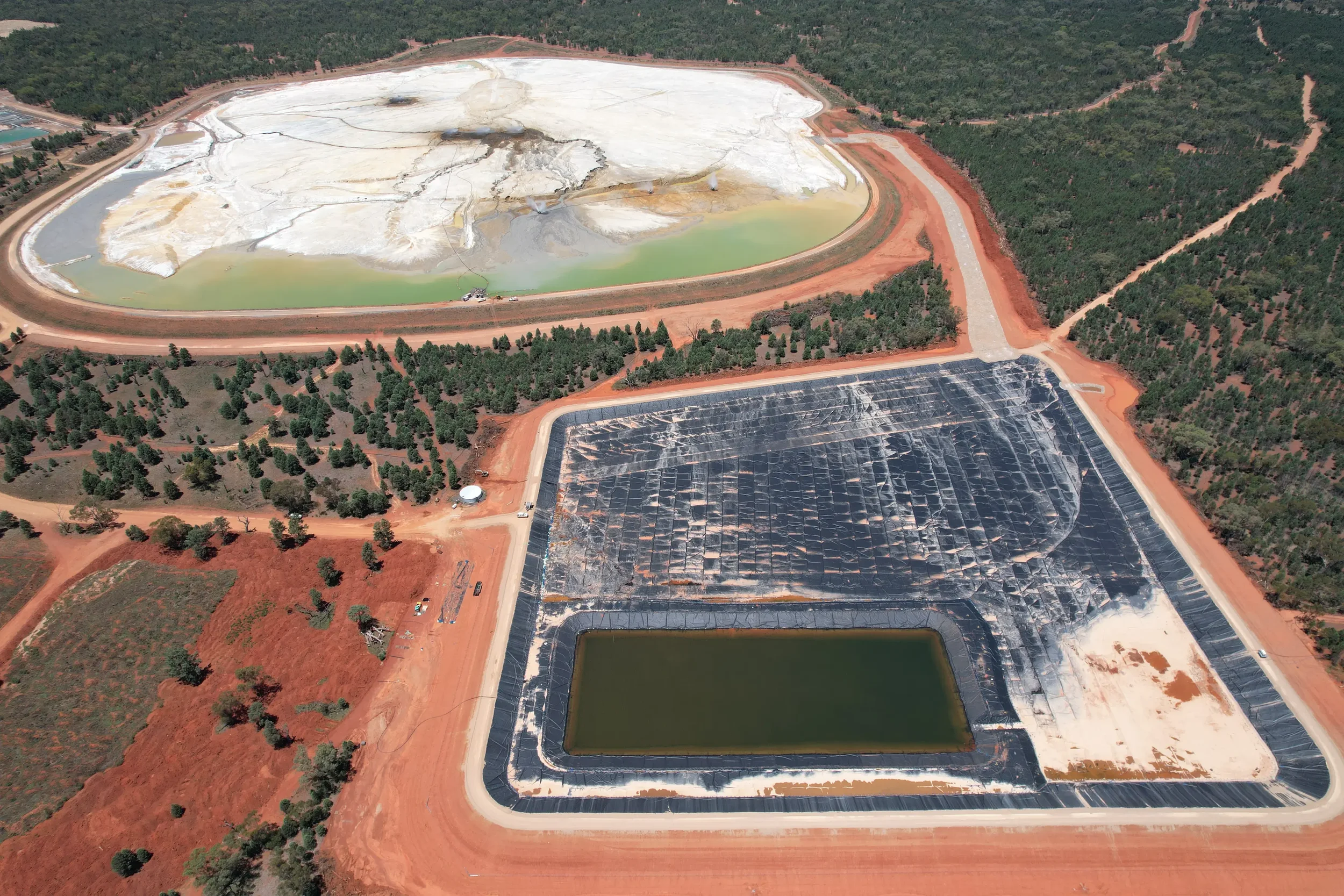 Aerial view of a mining operation showing large open-pit mines with white and brown material, holding ponds with green water, and surrounding roads and forested areas.