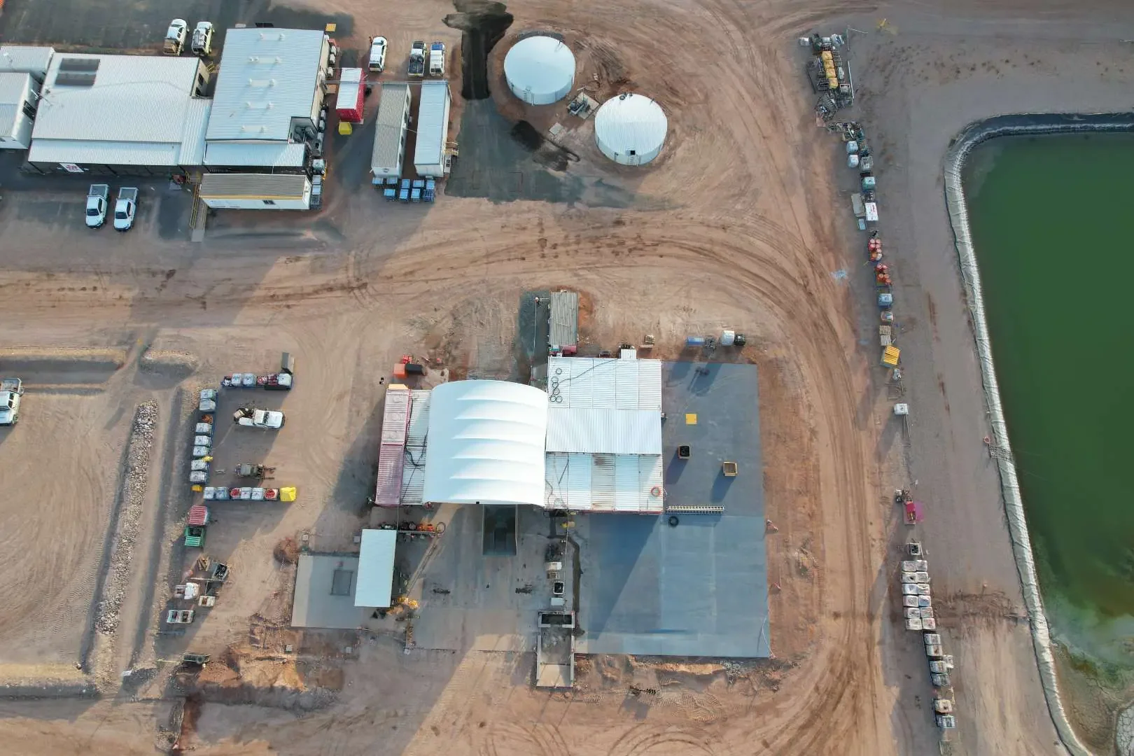 Aerial view of a construction site near a body of water with several buildings, storage tanks, and parked vehicles, including trucks and cars.