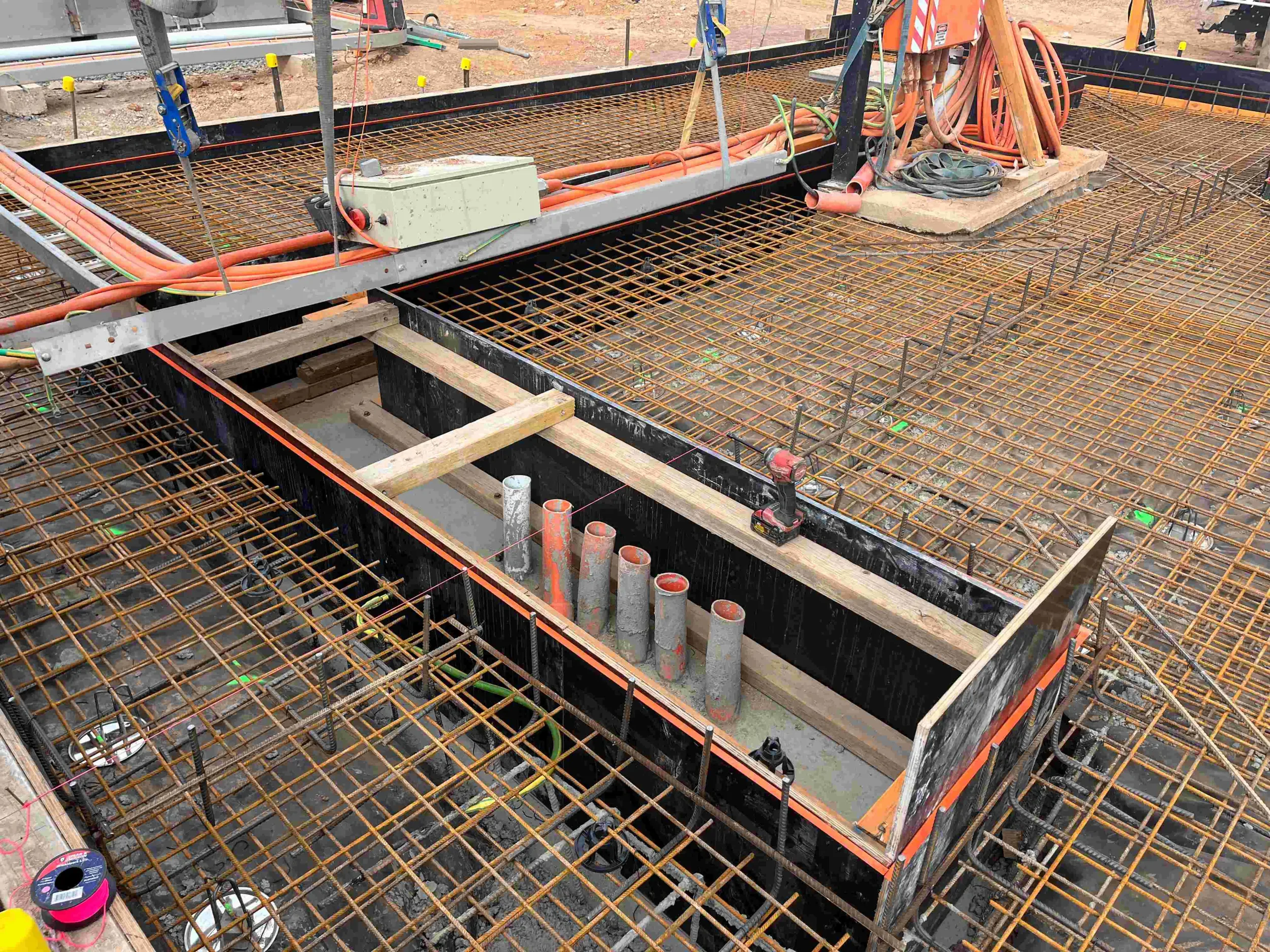 Construction site with steel rebar framework for concrete foundation, electrical and plumbing conduits, tools, and equipment for building a structure.