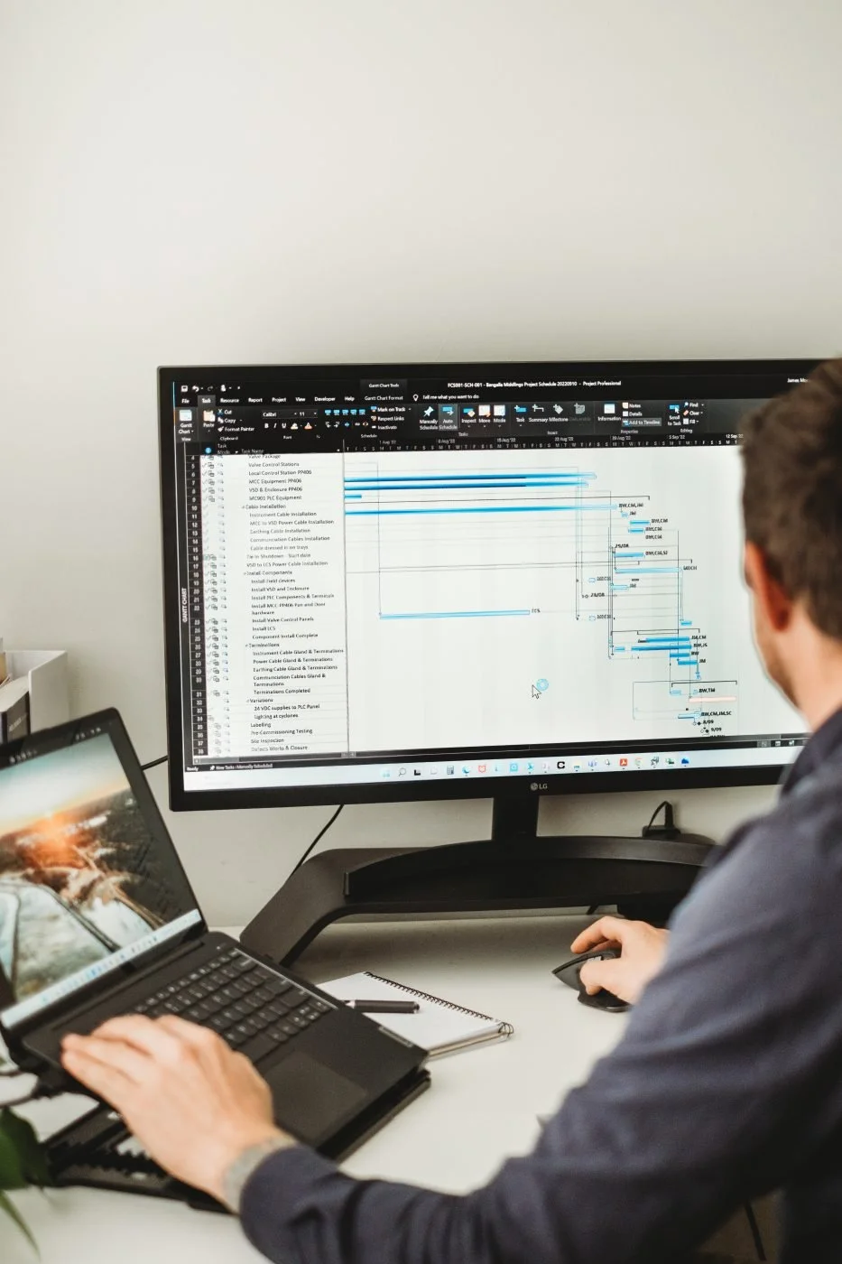 Person working at a desk with a laptop and a large computer monitor displaying a project management or Gantt chart.