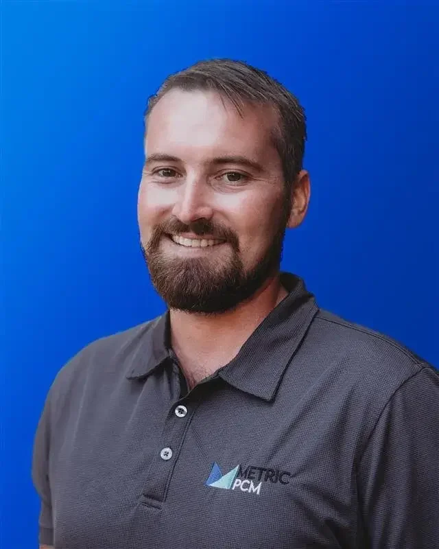 Portrait of a smiling man with short brown hair and a beard, wearing a dark gray polo shirt with a logo and the words 'Metric PCM' on it, standing against a solid blue background.