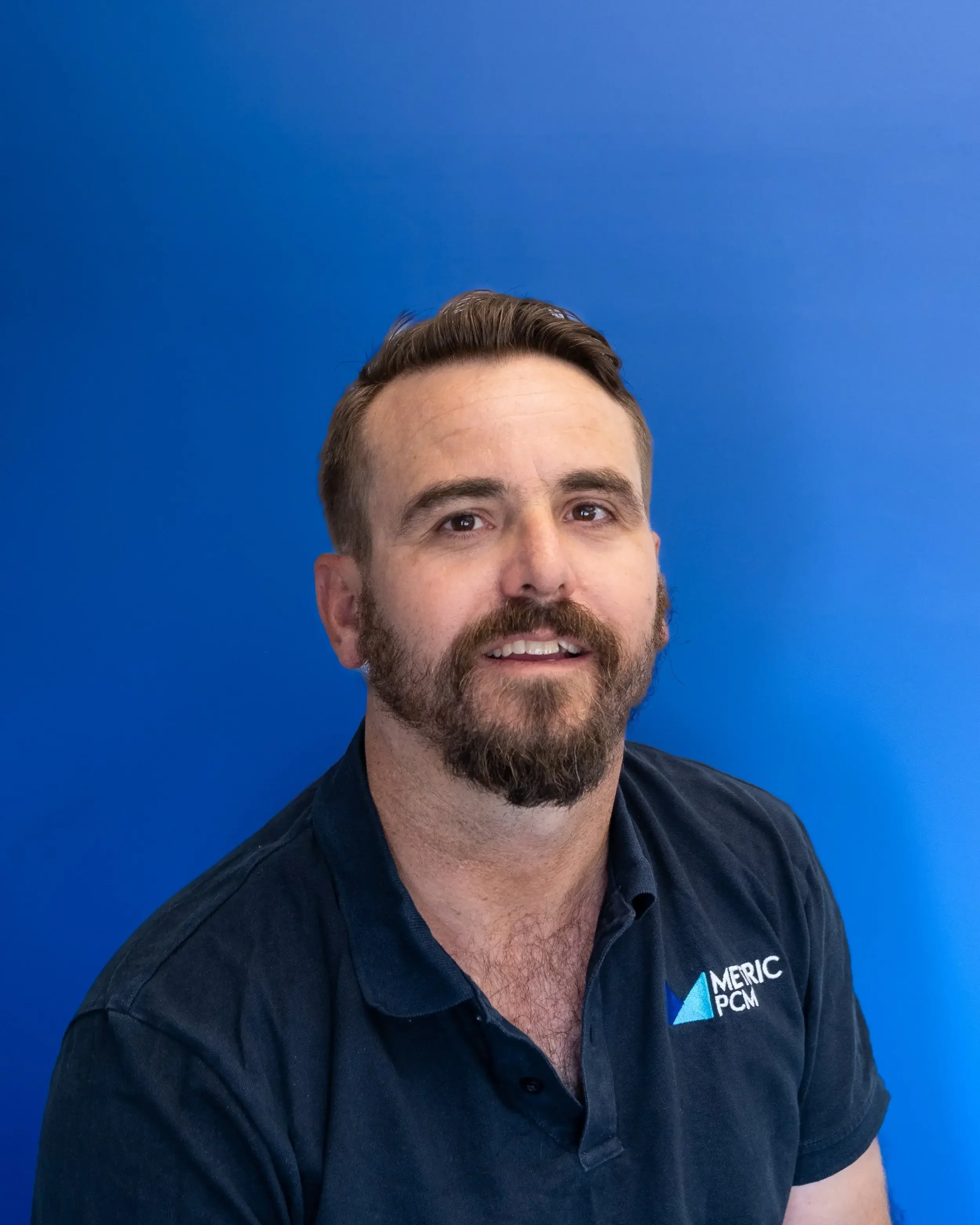 A man with short brown hair and a beard smiling, wearing a dark blue collared shirt with a logo that reads 'MERIC PCM,' against a solid blue background.