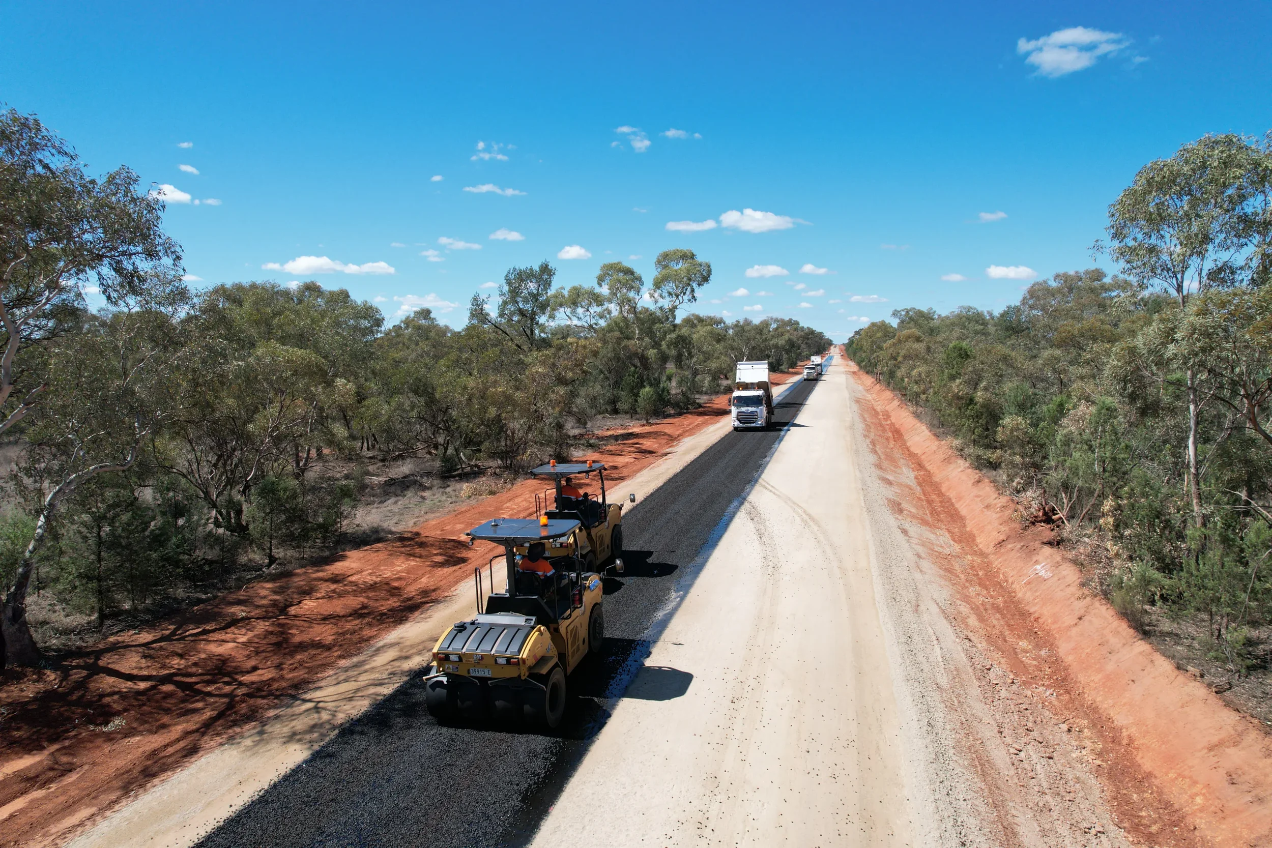 Construction vehicles laying asphalt on a rural road surrounded by sparse trees under a partly cloudy blue sky.