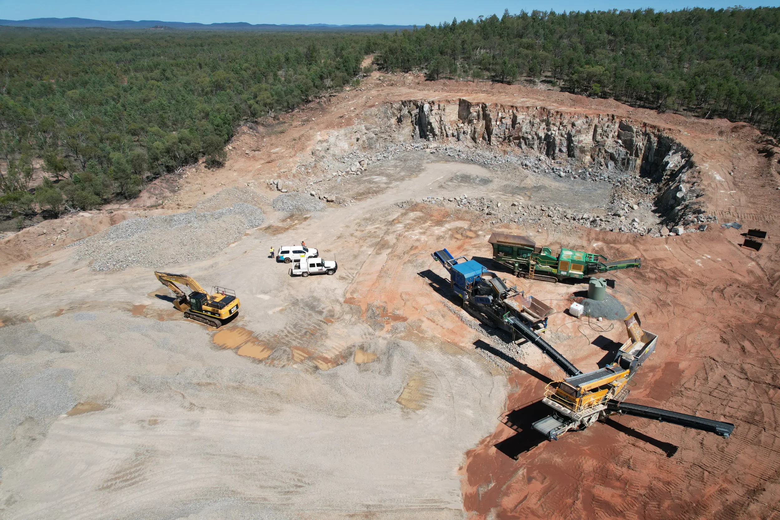 An aerial view of a quarry site with heavy machinery, trucks, and thick forest surrounding it, with mountains in the distance.