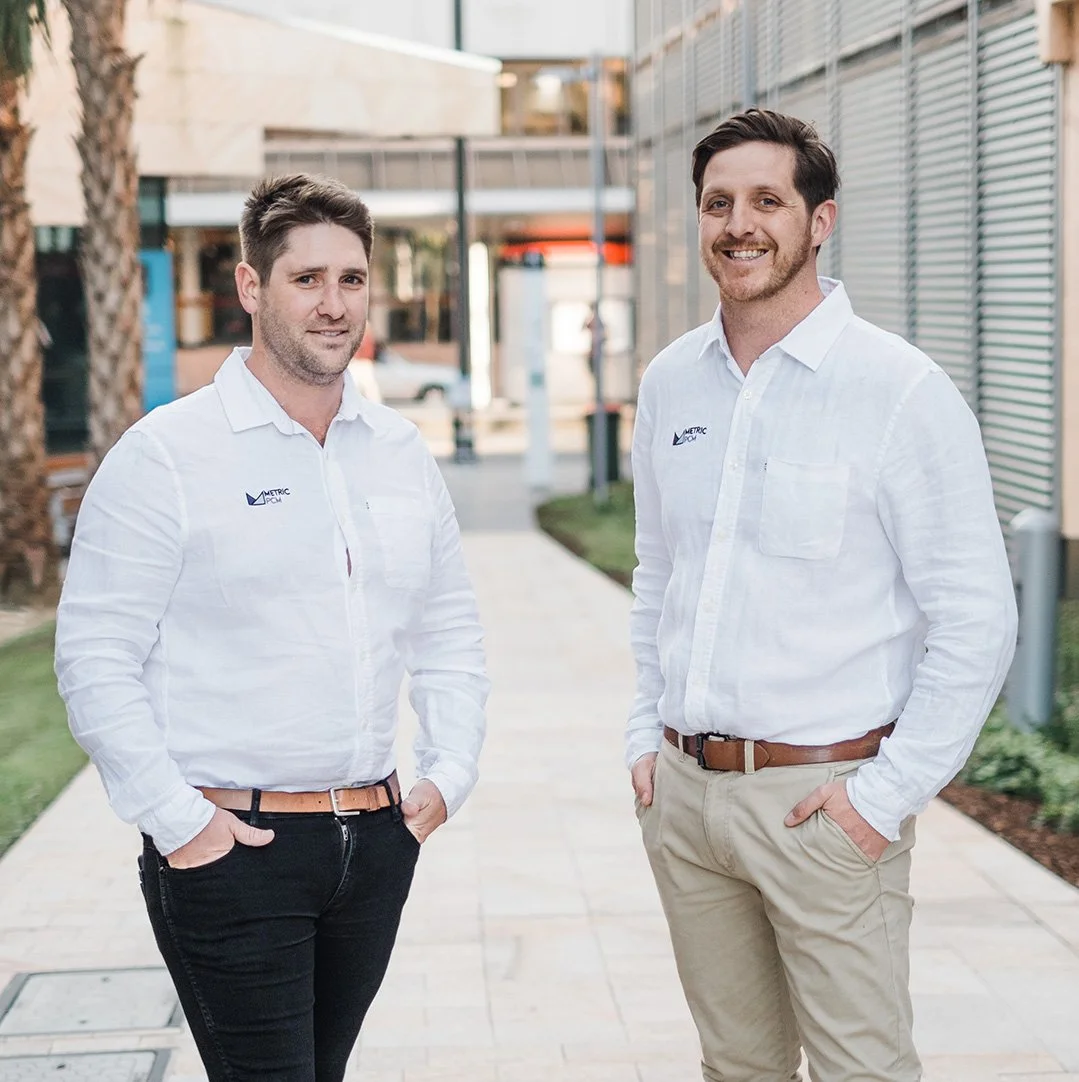 Two men standing outdoors in casual business attire, smiling, in an urban setting with buildings, trees, and a sidewalk.