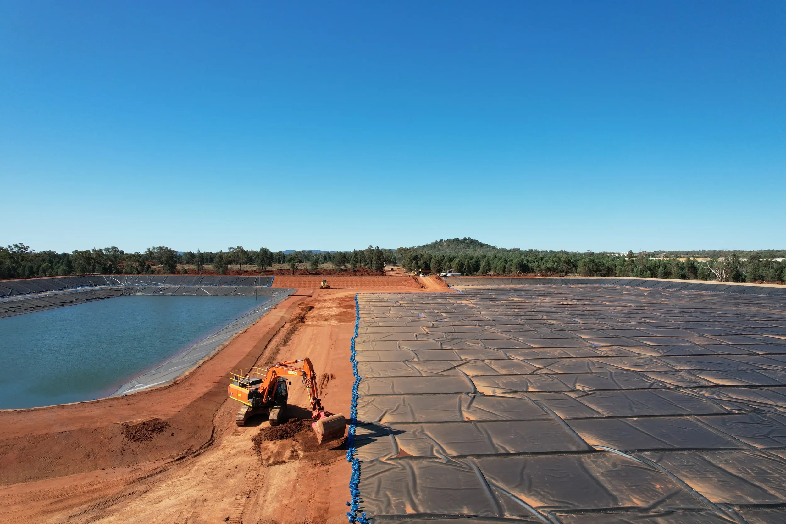 Construction site with a large body of water lined with black plastic and blue piping, a small excavator in the foreground, and dirt roads extending into a landscape of trees and hills under a clear blue sky.