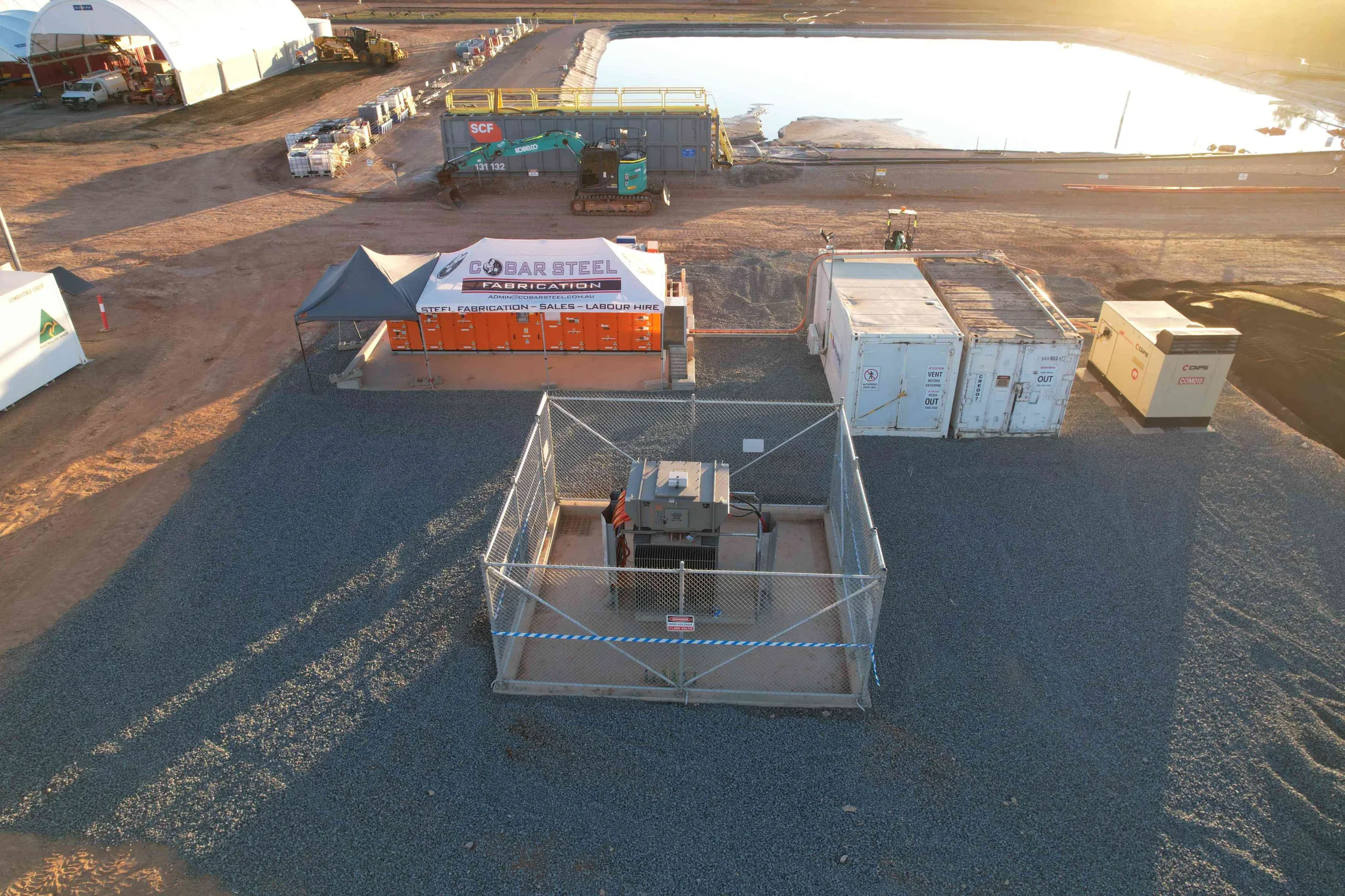 Construction site with machinery and containers near a body of water in the background, a tent with company branding, and various equipment and supplies.